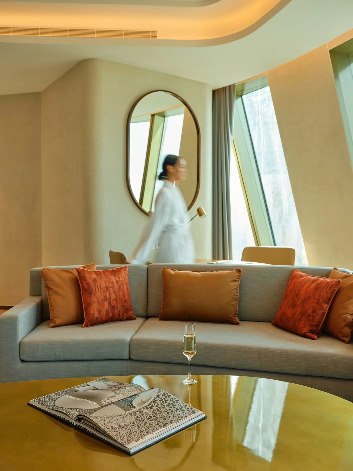 A modern hotel suite with a gray sofa with orange pillows, a glass of champagne on a wooden coffee table, an open magazine, a woman in white walking in a blurred motion, a large oval mirror on the wall, and large windows with angled green frames and curtains.