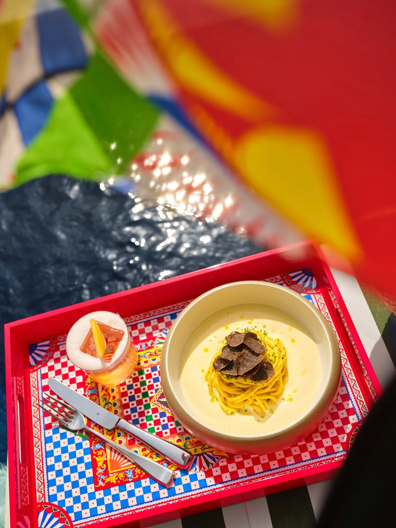 A tray holding a bowl of spaghetti with black truffle slices on top, a small cup with a pink cocktail garnished with a lemon wedge, a fork and knife, all on a colorful patterned tray.