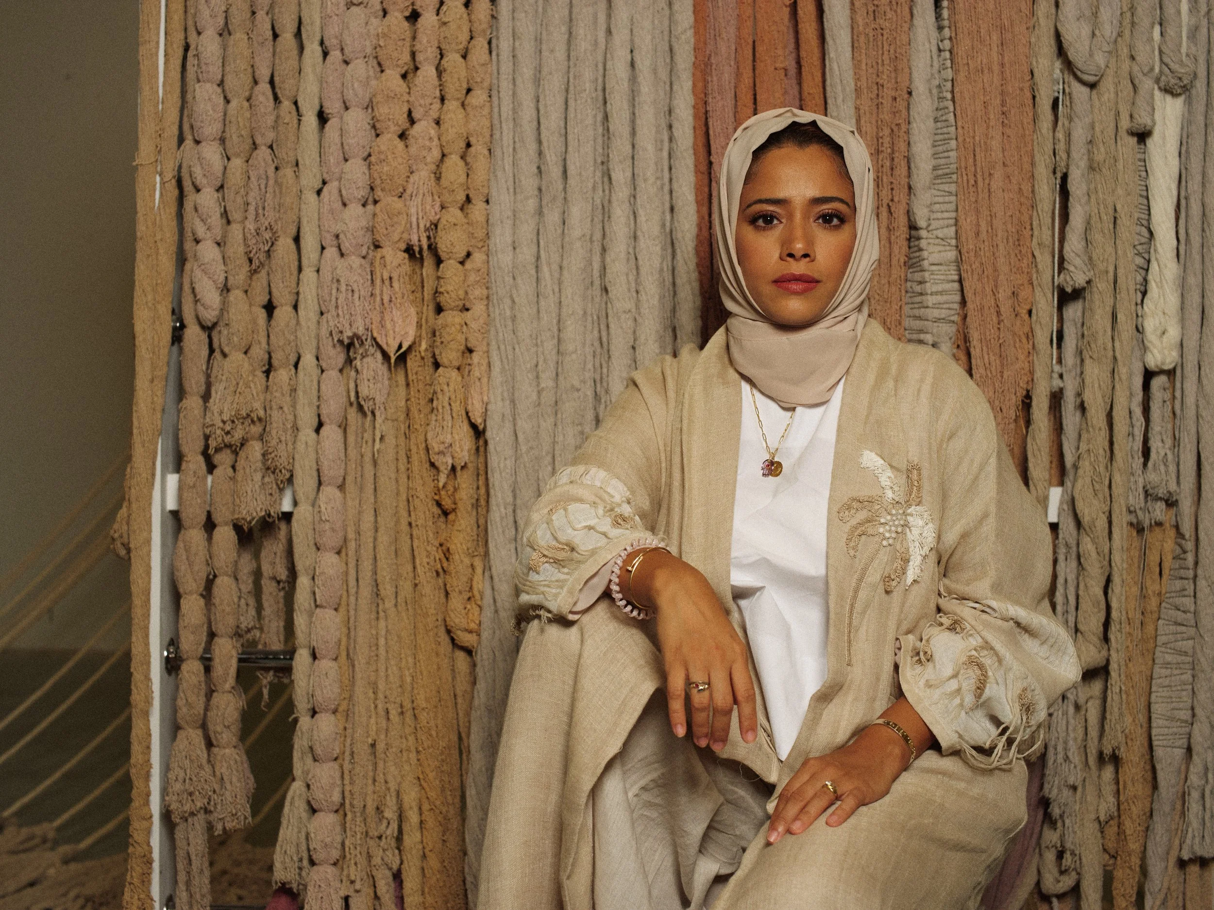 A woman sitting in front of a textured backdrop of woven fabric and rope, wearing a light beige headscarf, beige jacket with embroidered designs, a white shirt, and jewelry including necklaces, rings, and bracelets.