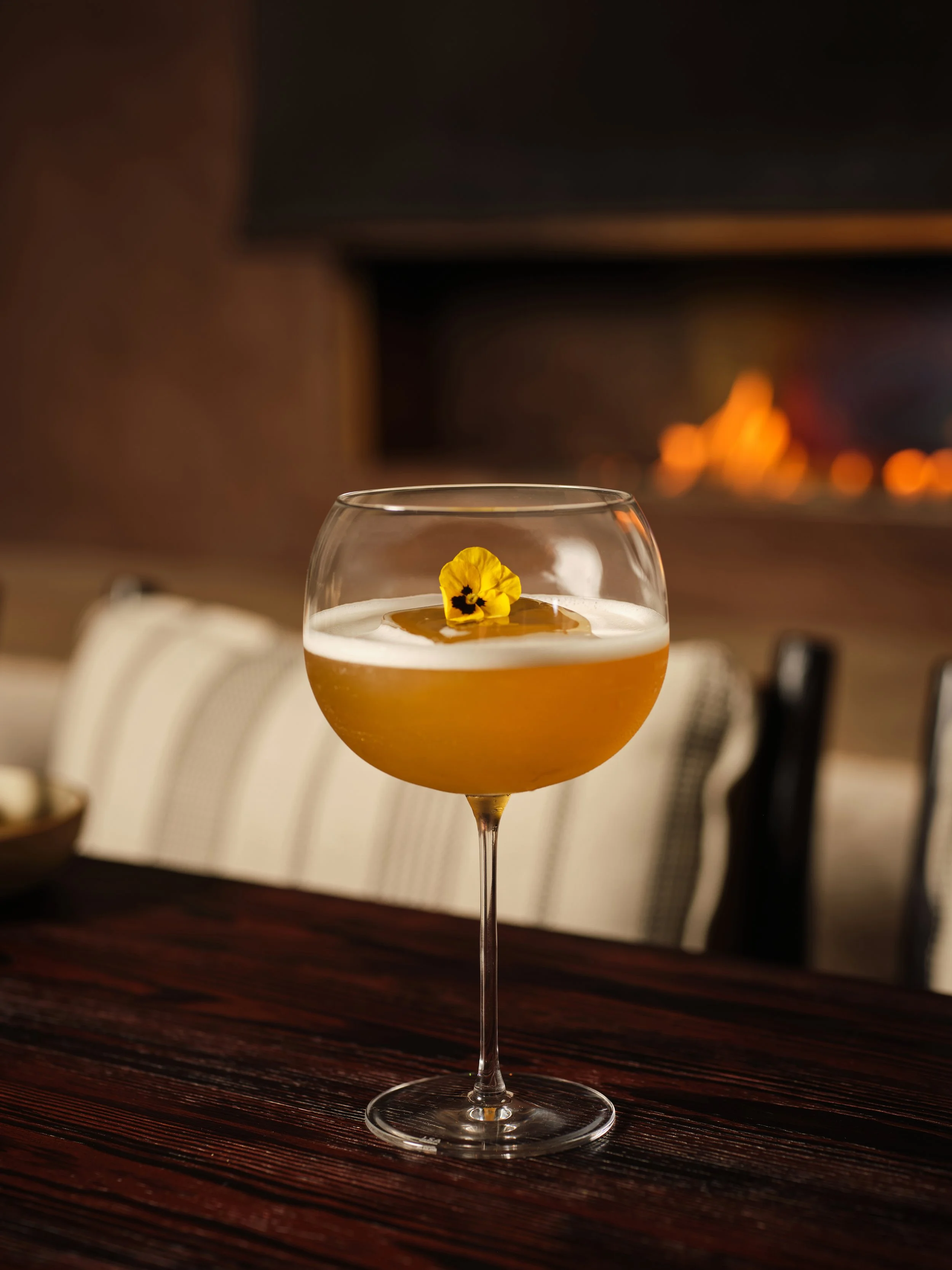 A cocktail glass with a yellow-orange drink topped with a small yellow flower and a brown garnish, set on a dark wooden table in front of a brick fireplace with a fire burning.