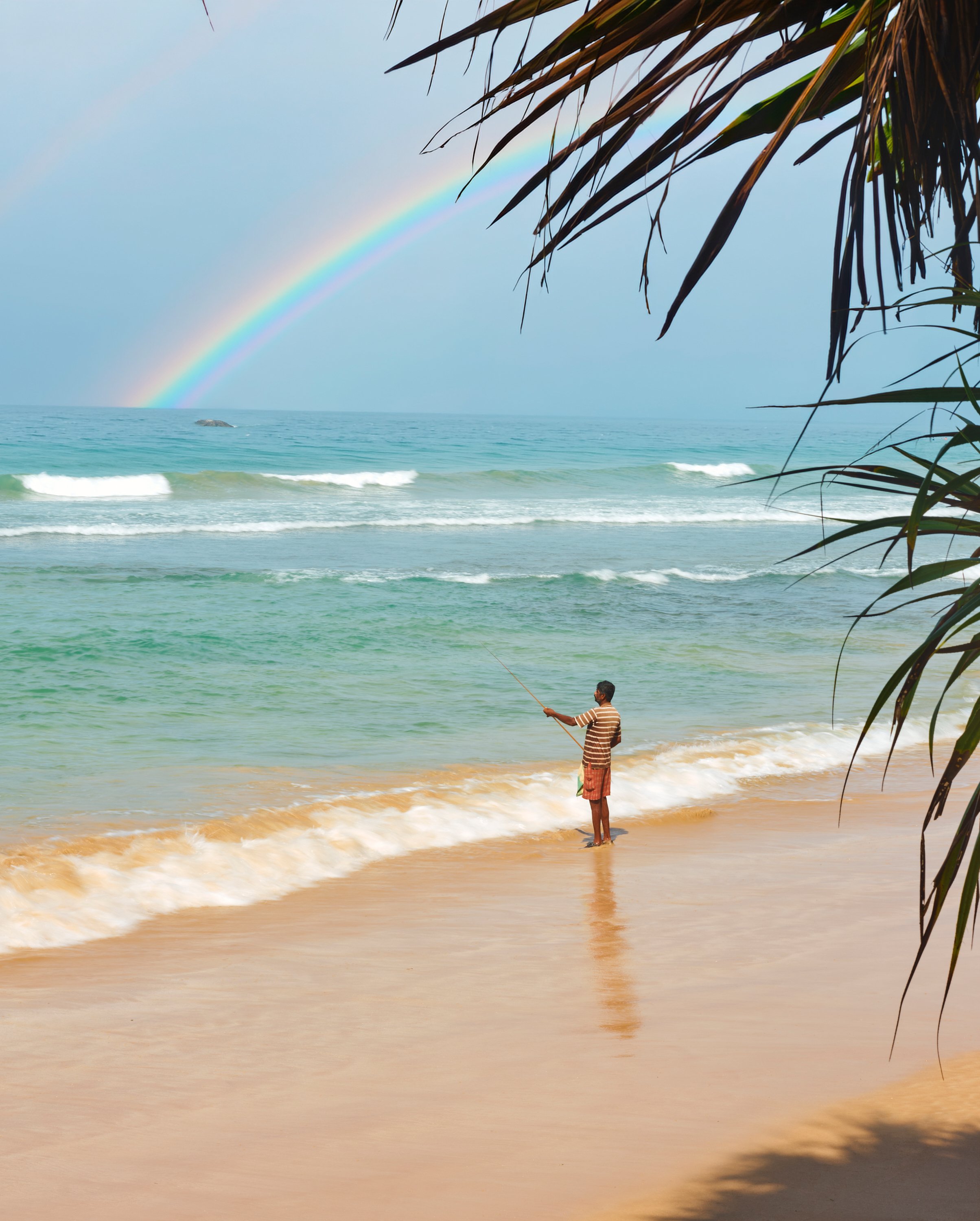 A man standing on a sandy beach near the water, fishing with a fishing rod, with a rainbow in the cloudy sky and palm leaves framing the scene.