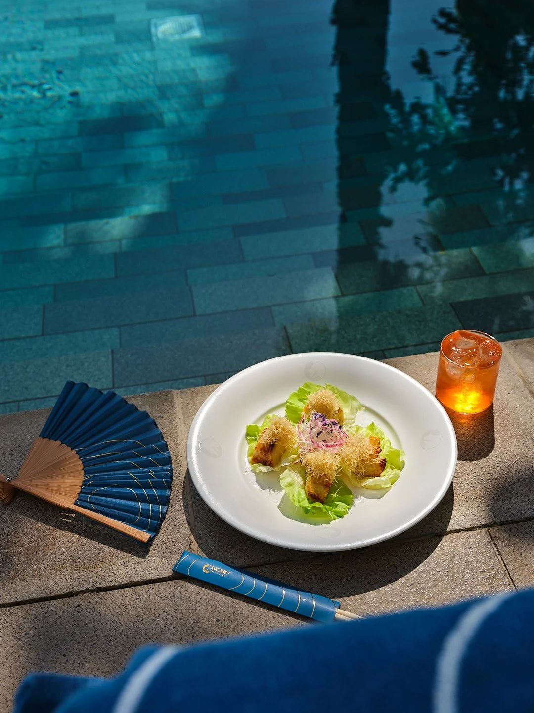 A plate with pieces of sushi topped with shredded garnish, on a bed of lettuce, placed on a stone ledge by a swimming pool. To the right of the plate is a small glass of amber-colored drink with ice. On the left side of the image, a blue and brown fan and a blue chopstick are visible.