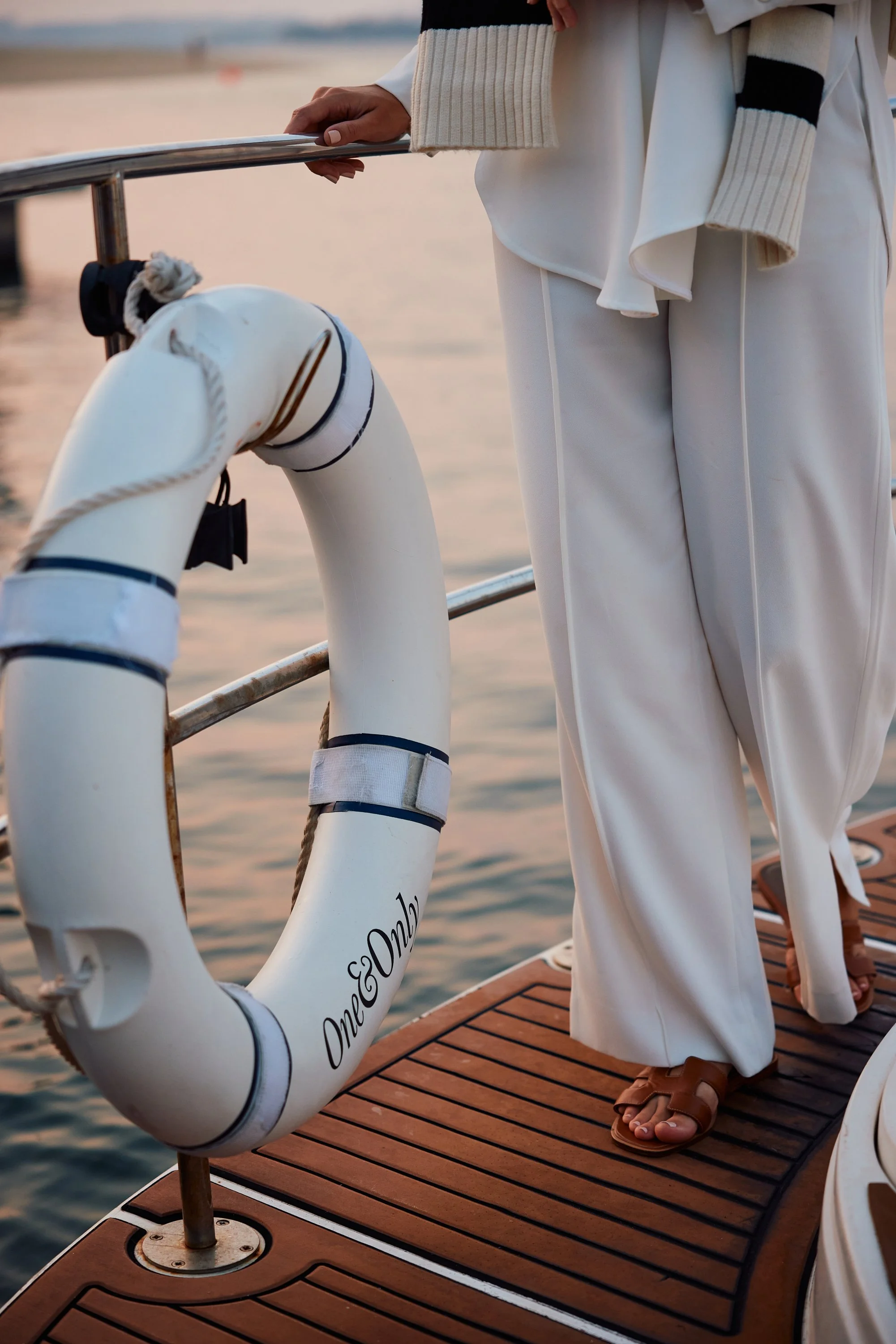 Person standing on a boat deck near the water, with a life preserver attached to the boat railing, dressed in beige and white clothing with sandals.