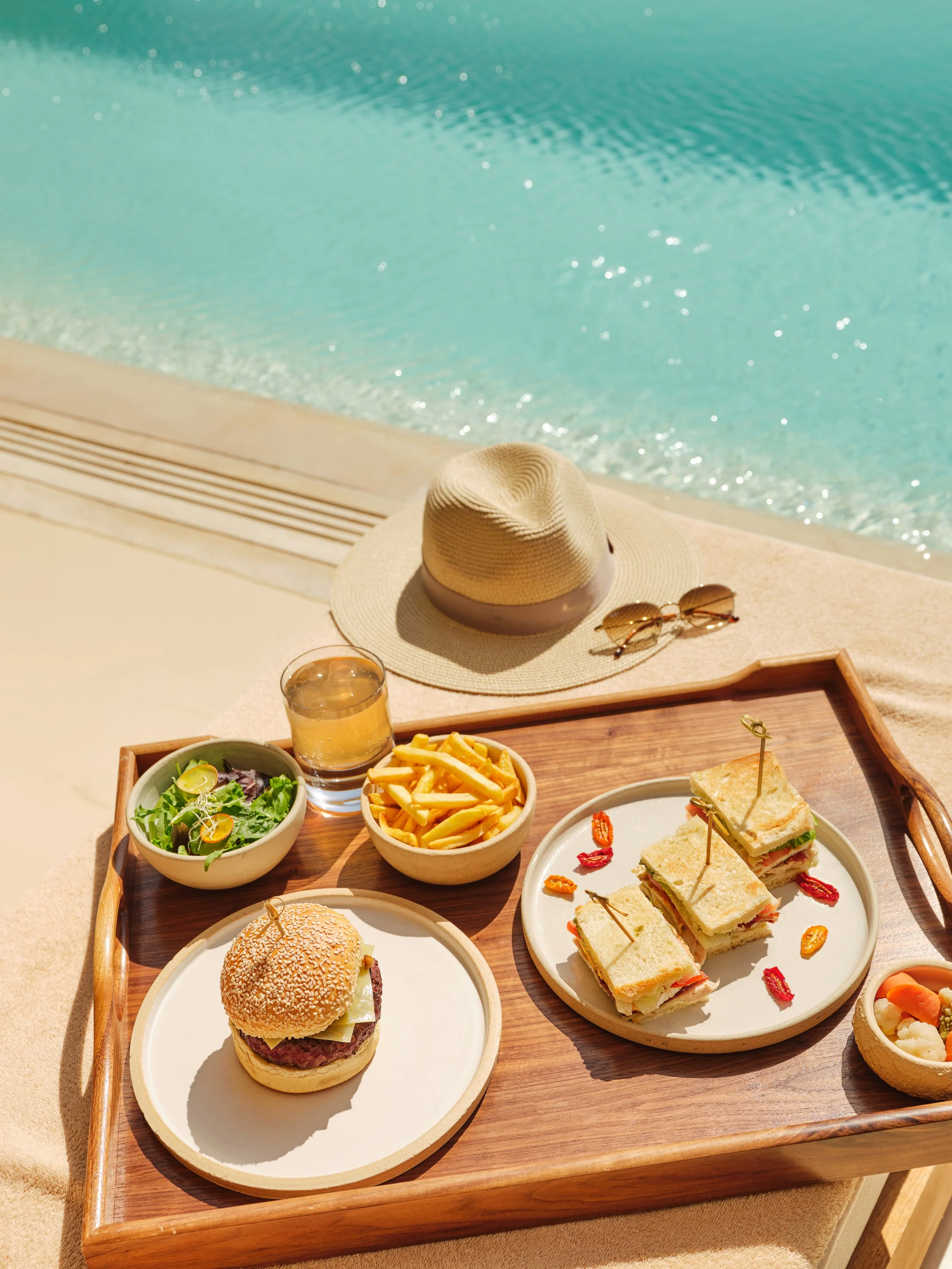 Tray with a small burger, fries, salad, sandwich, and drinks by a poolside, with a sun hat and sunglasses nearby.