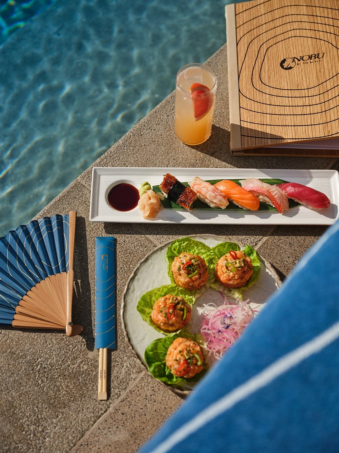 Sushi platter with assorted sushi, soy sauce, wasabi, and pickled ginger, along with a drink, fan, and wooden menu on a poolside table
