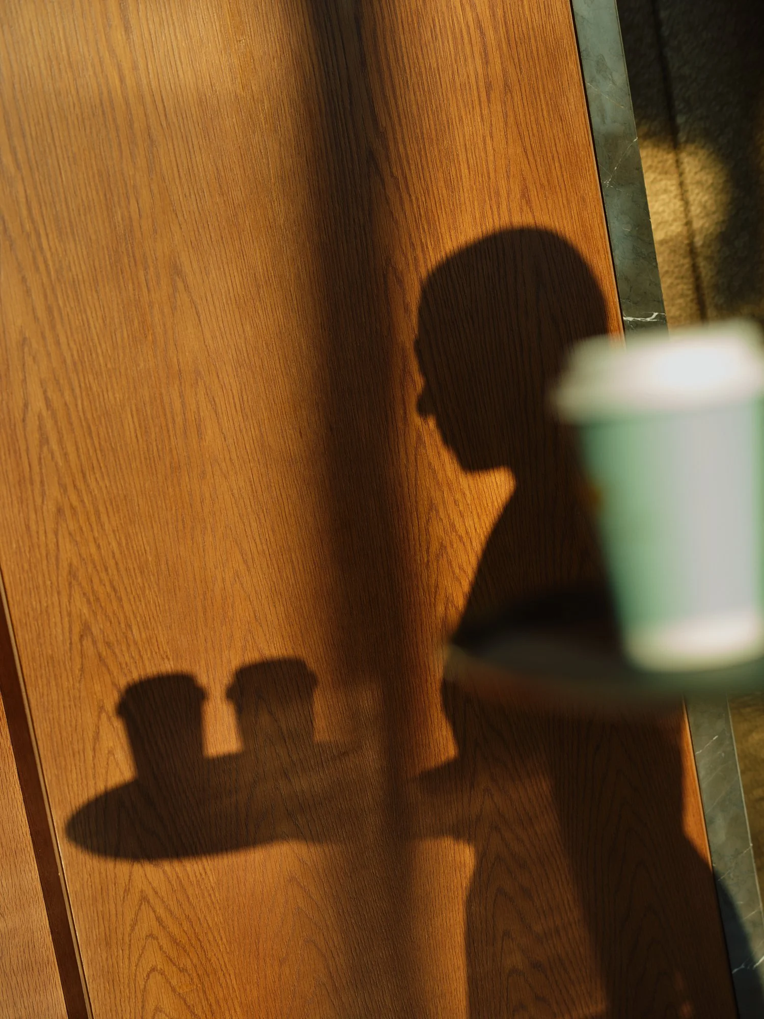 Shadows of three people and a cup on a wooden surface with a green container in the foreground.