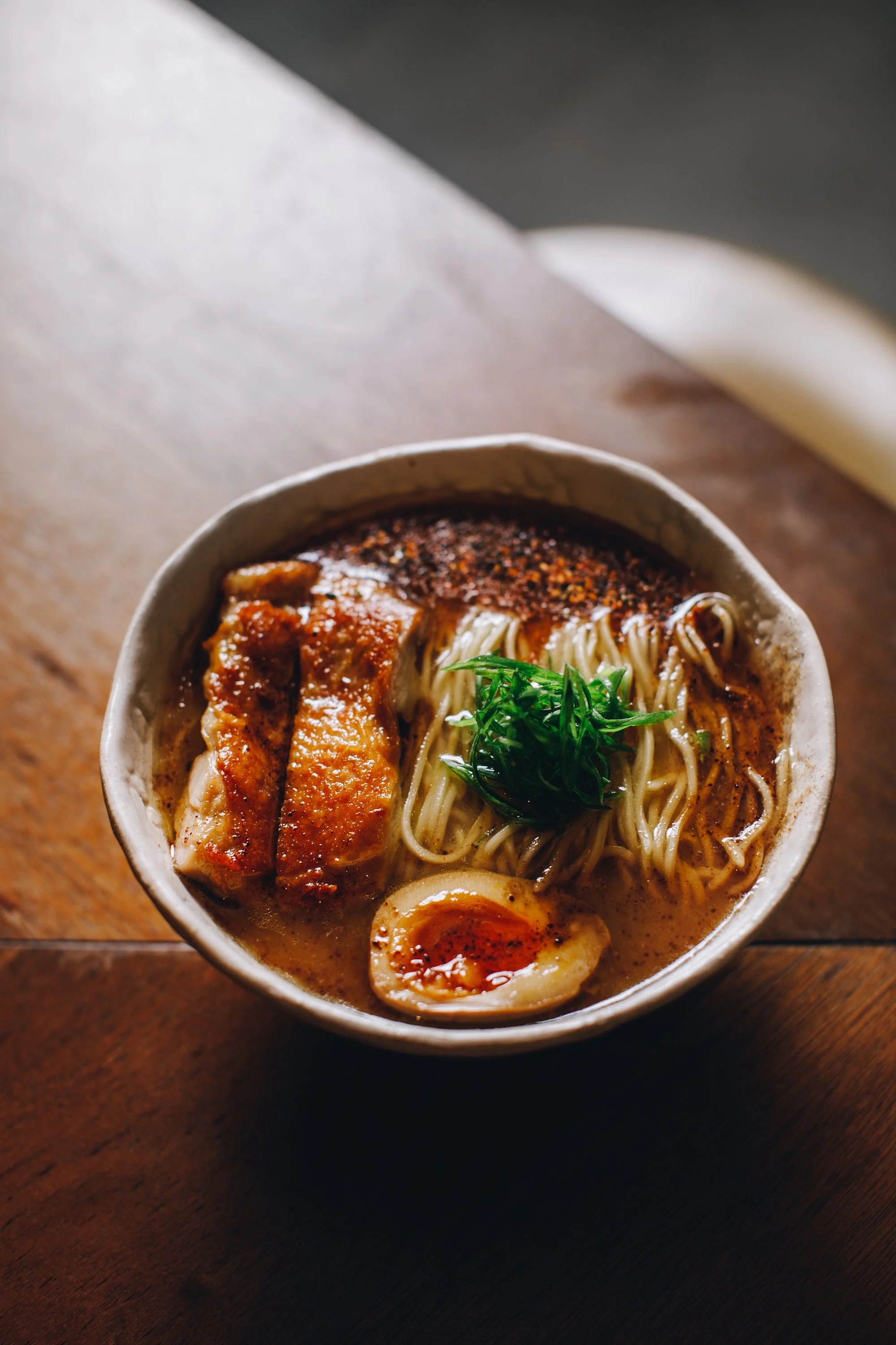 A bowl of ramen noodles topped with sliced pork, a soft boiled egg, green herbs, and spicy seasoning on top, served on a wooden table.