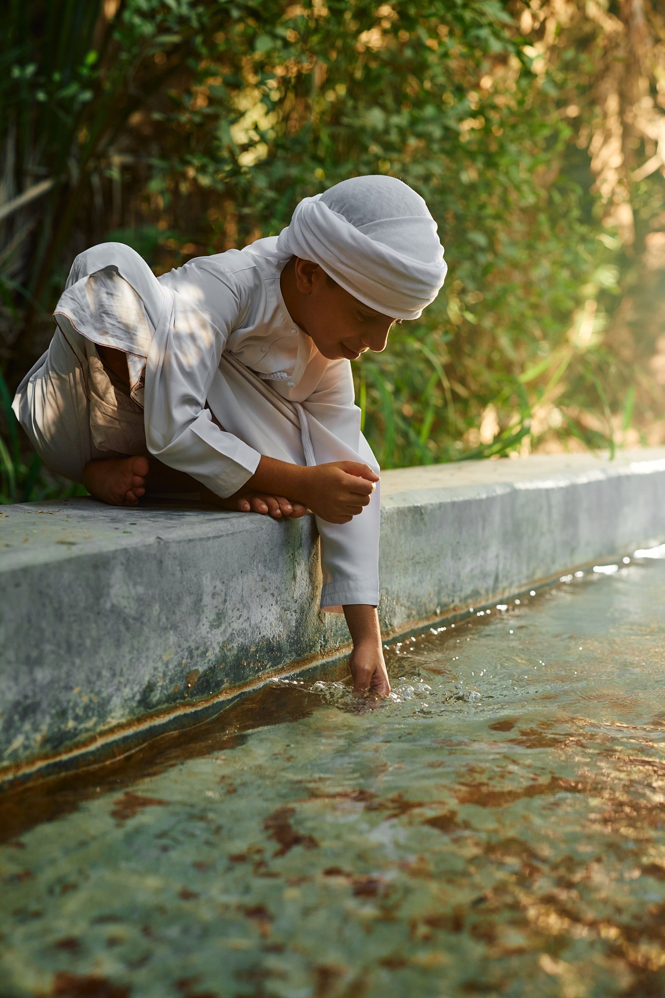 A young person dressed in white clothes and a white head covering kneeling on a concrete ledge by a waterbody, reaching into the water with their hand in a lush green area during daylight.