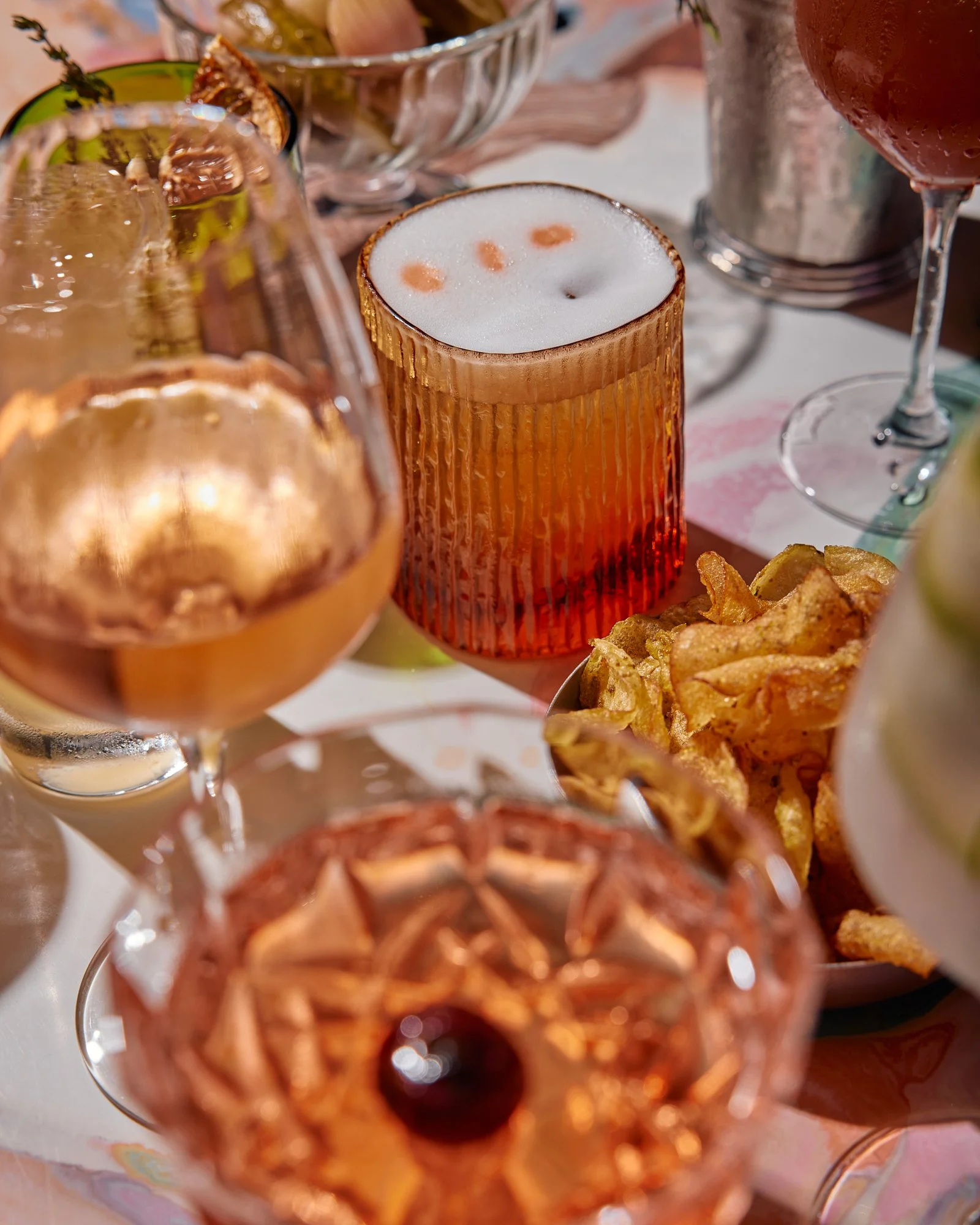 Various cocktails, chips, and snacks on a table, including a drink with foam and bubbles, and another with a cherry on top.