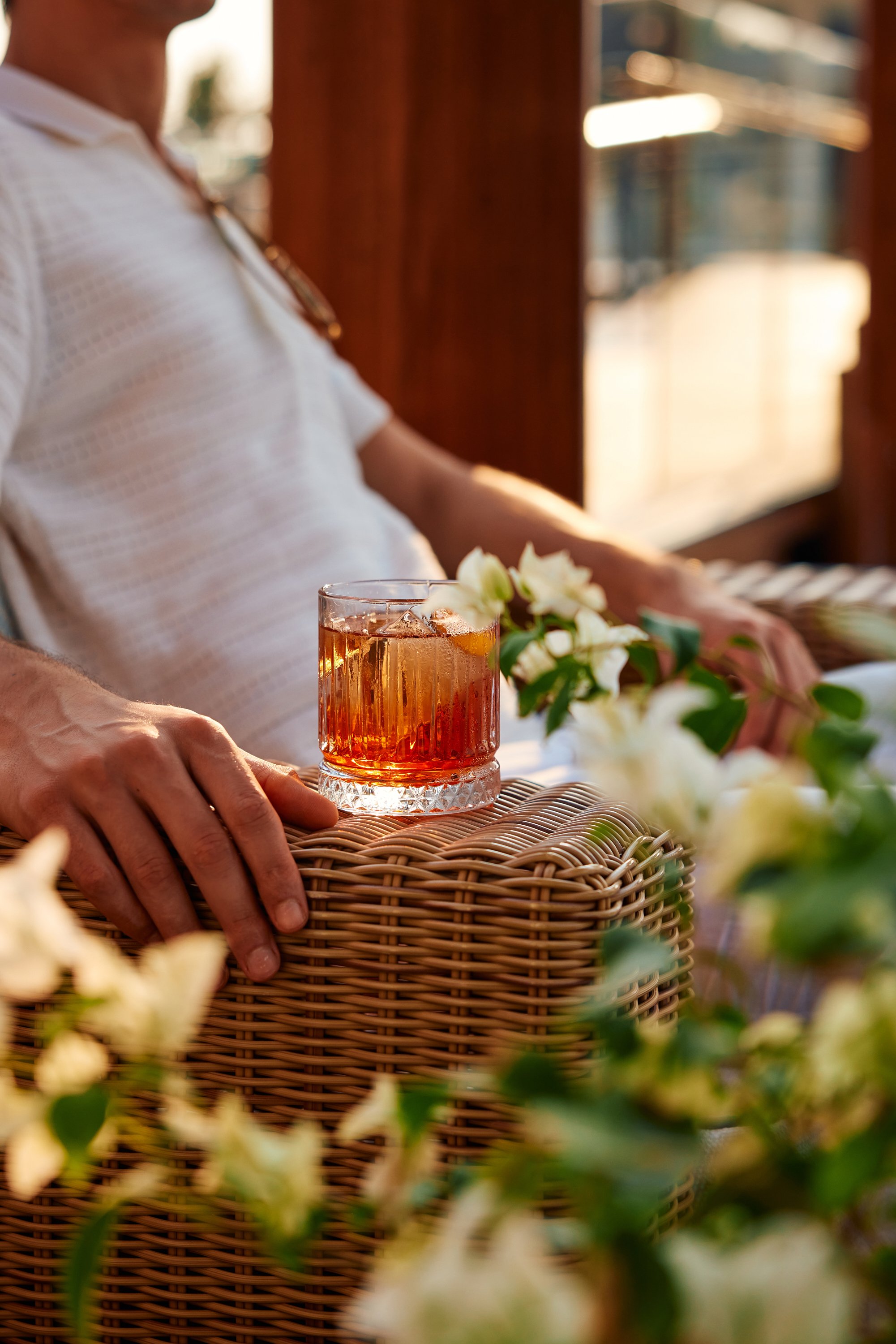 Person resting at a table with a wicker surface, holding a glass of iced tea with ice cubes, surrounded by white flowers, in a sunlit setting with wooden walls and windows.