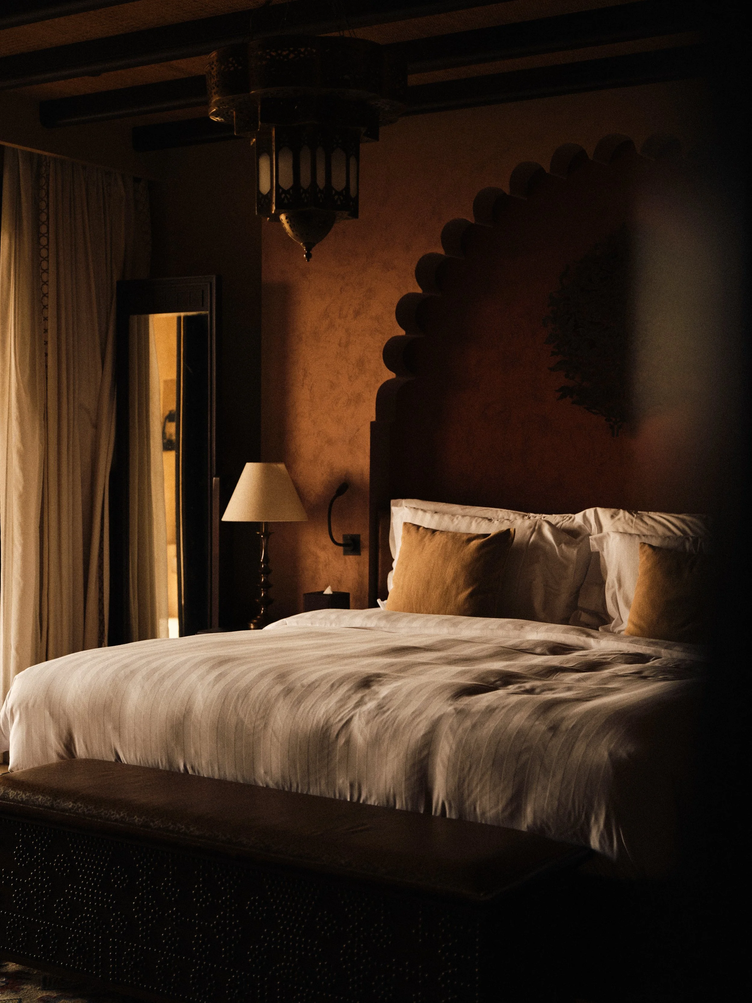 A cozy hotel room with a large bed, beige pillows, and white linens, illuminated softly with a bedside lamp, featuring curtains and a mirror on the wall.