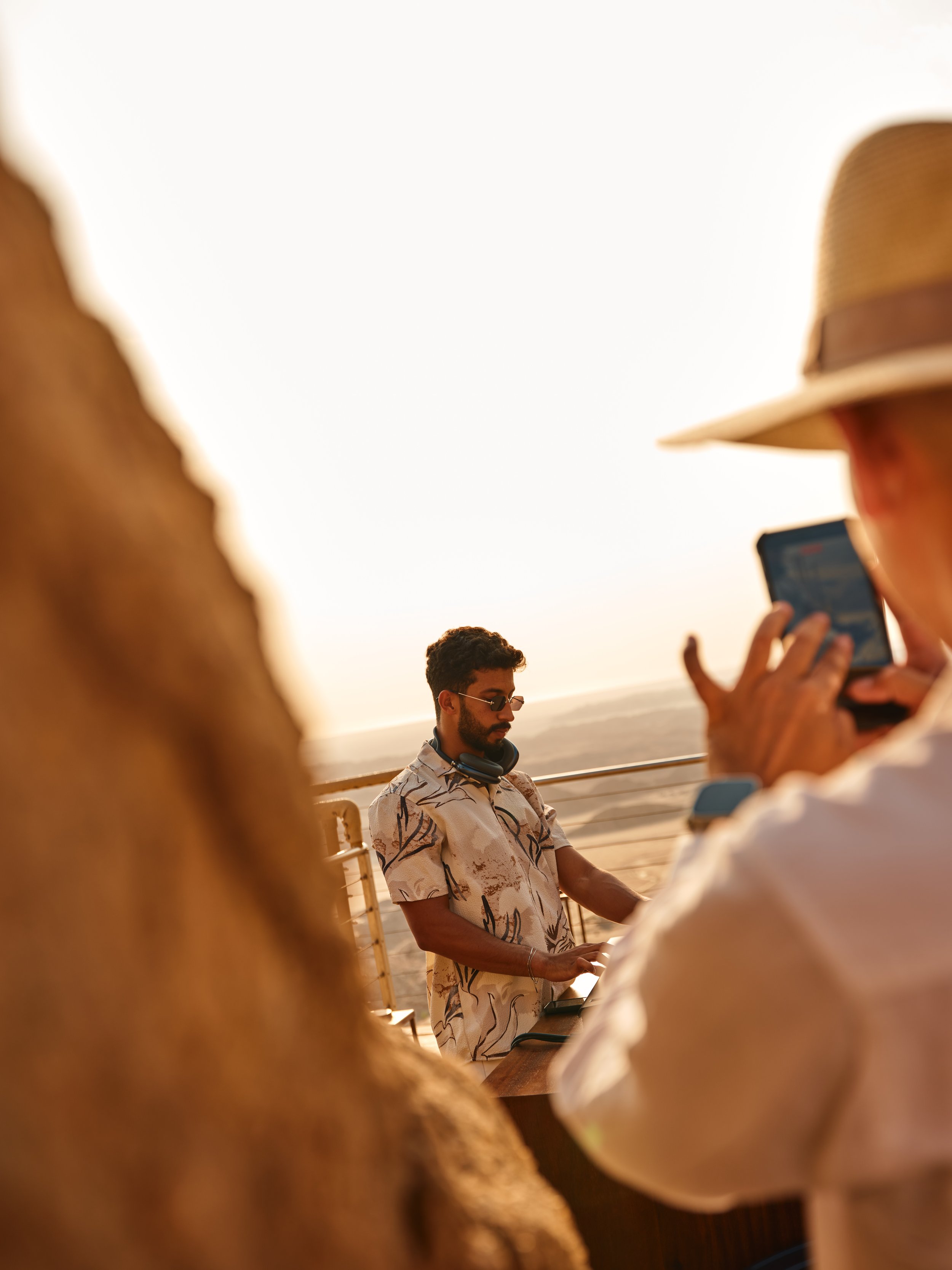 A man with headphones around his neck, wearing sunglasses and a patterned shirt, standing at a DJ station outdoors during sunset, with two people in the foreground, one of whom is taking a photo with a smartphone.