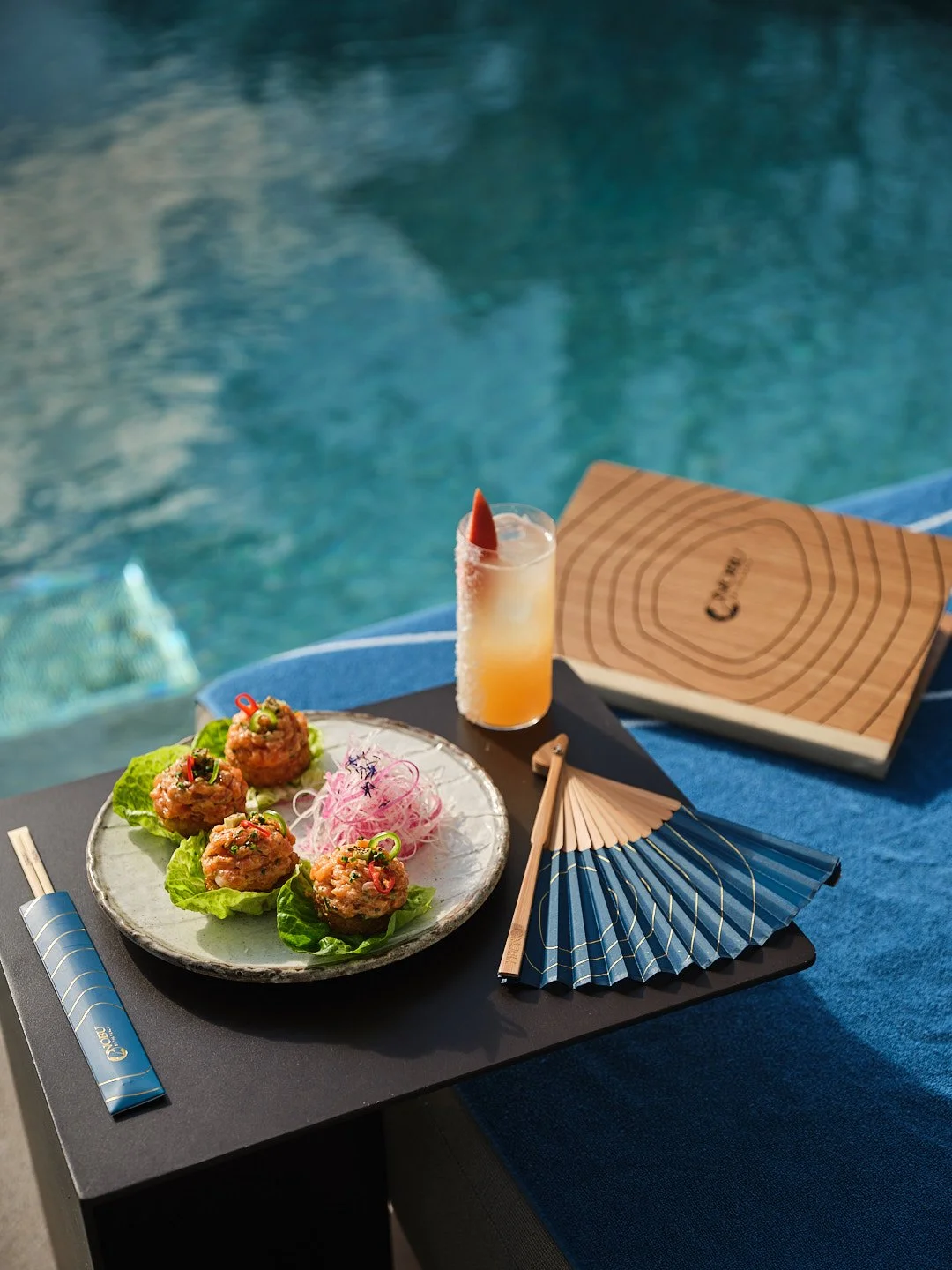 Plate of Asian meatball appetizers garnished with chopped herbs and sliced peppers, with pink shredded vegetables on the side, placed on a black table near a swimming pool.