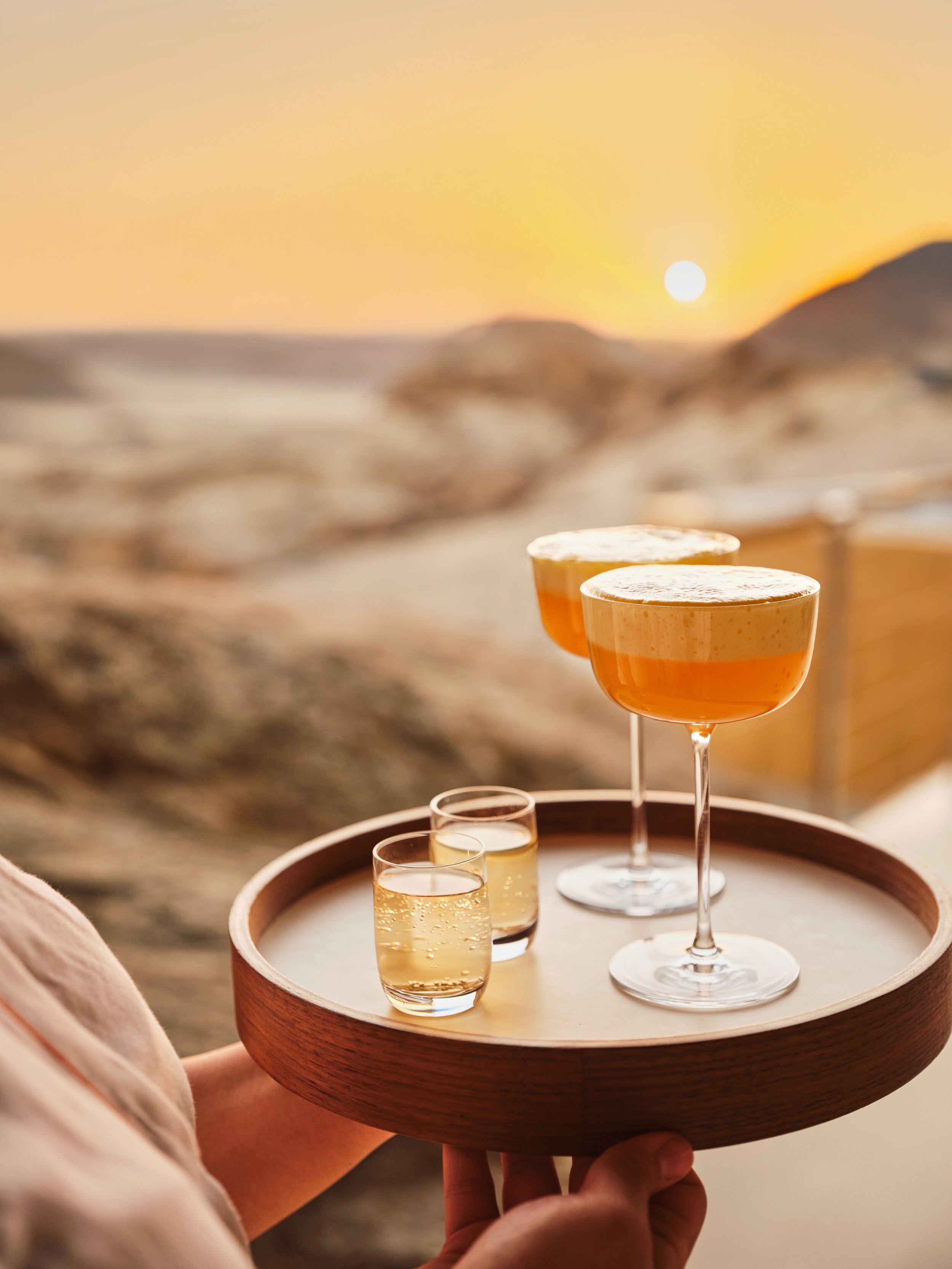 A person holding a round wooden tray with two cocktail glasses and two shot glasses, with a desert landscape and setting sun in the background.