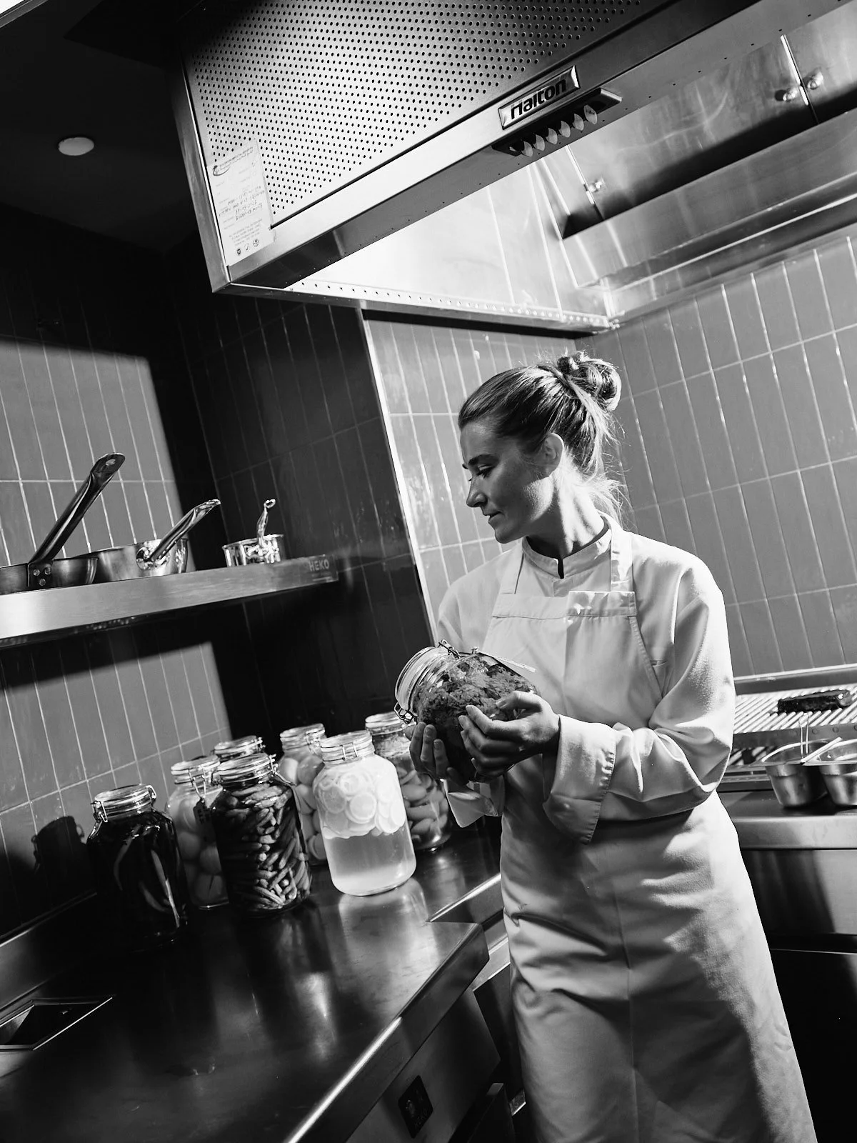 A woman in a chef's coat and apron in a commercial kitchen, examining a jar of preserved food, surrounded by jars of preserved fruits or vegetables on the counter.
