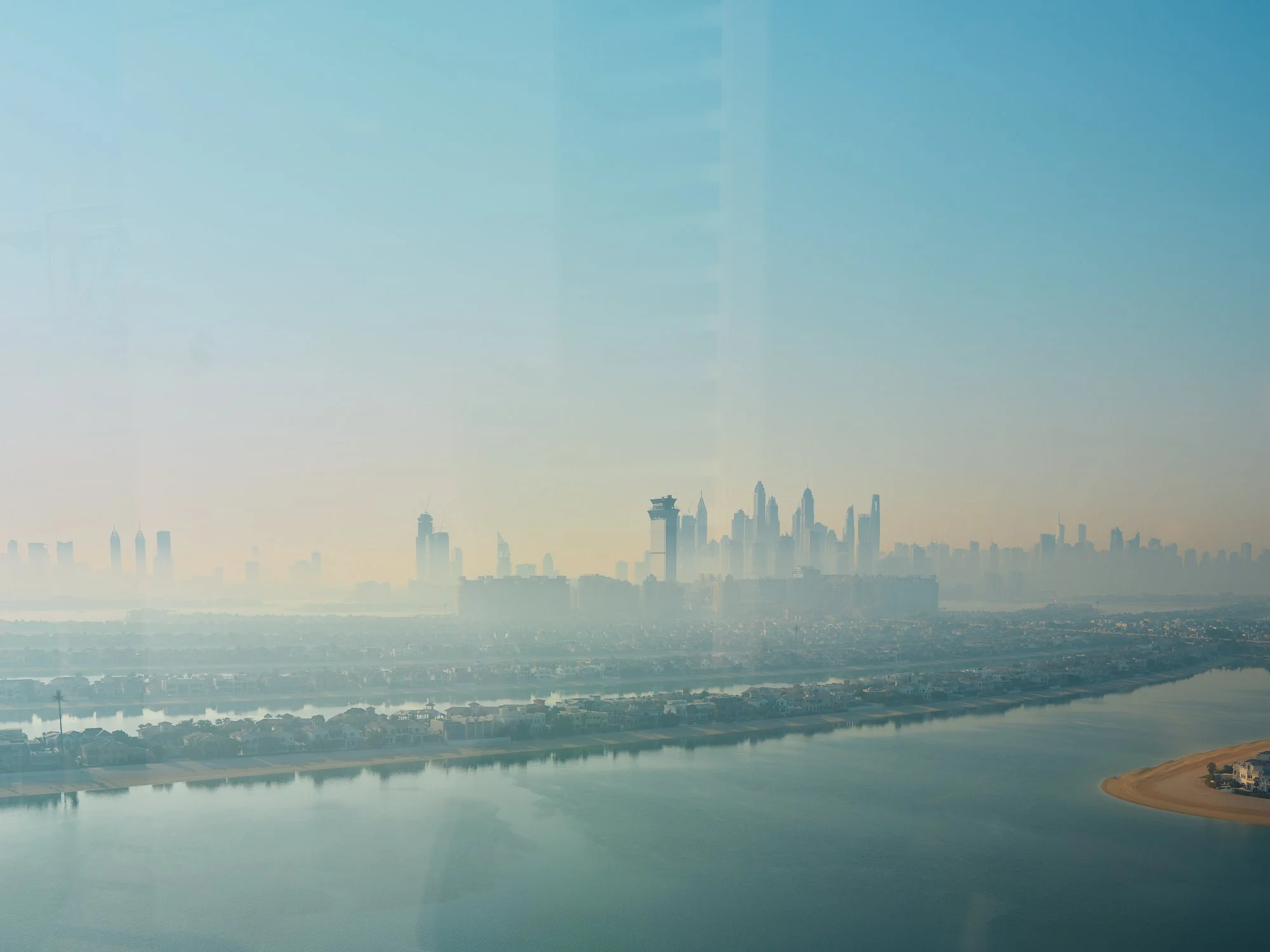 A city skyline with tall skyscrapers and buildings seen through a hazy, possibly foggy or polluted atmosphere, with a body of water in the foreground and a distant shoreline.