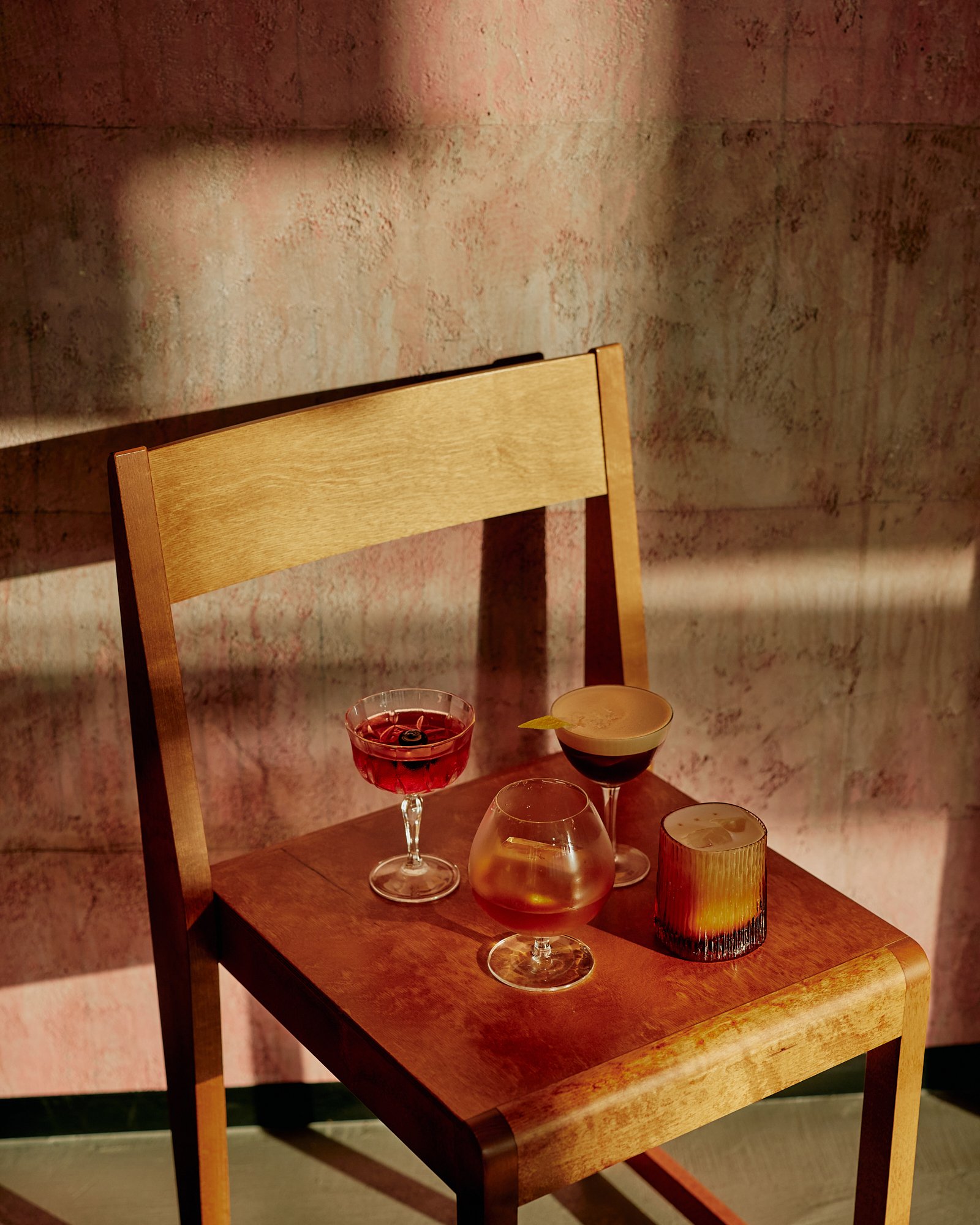 Four cocktails on a wooden chair in front of a textured wall with warm lighting.