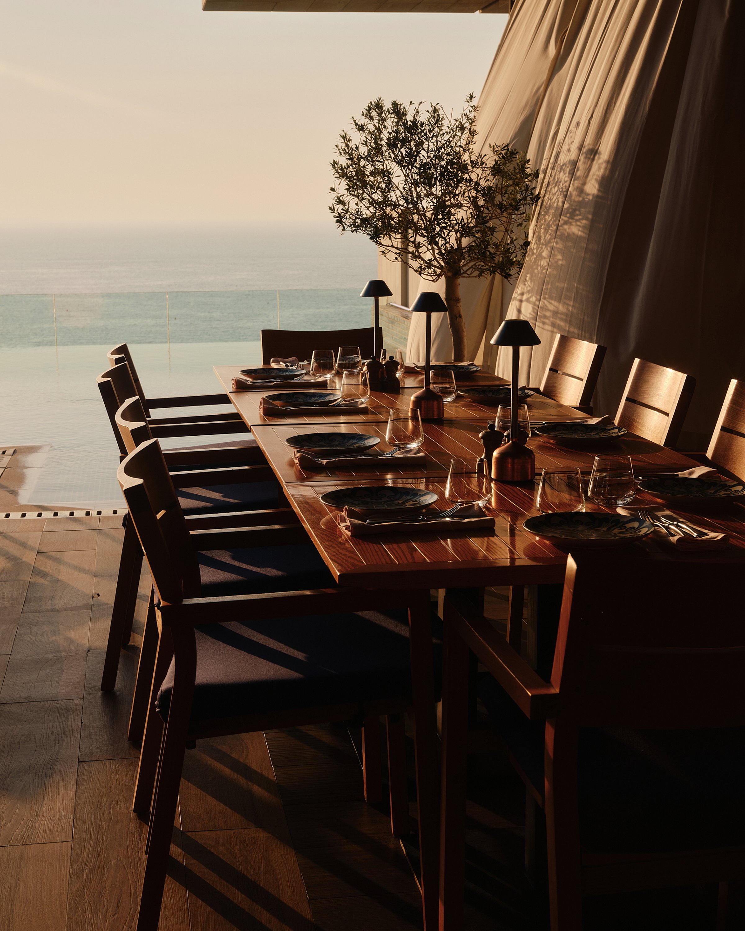 A dining table set with plates, glasses, and utensils, with a view of the ocean in the background. There are chairs around the table, large curtains, and a potted tree inside the room. The scene is illuminated by warm sunlight.