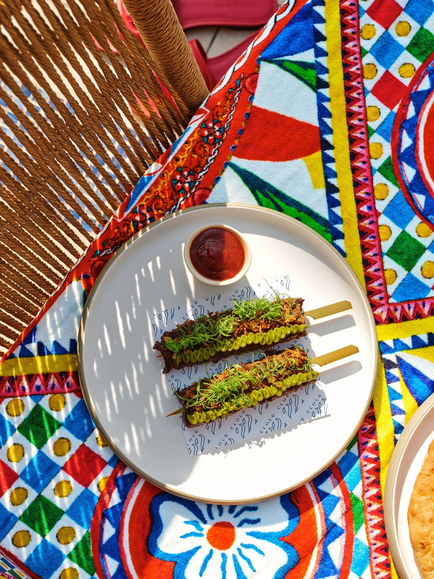 Two skewers of grilled meat topped with microgreens and mustard, served with a small cup of dipping sauce on a white plate, placed on colorful embroidered tablecloth, with a wooden woven chair beside.