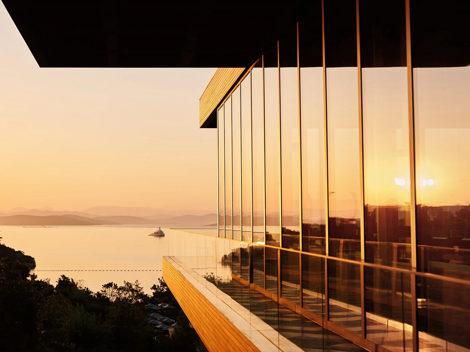 Sunset view of a modern building with glass windows reflecting the sunset, overlooking a body of water with a boat, trees in the foreground, and distant mountains.