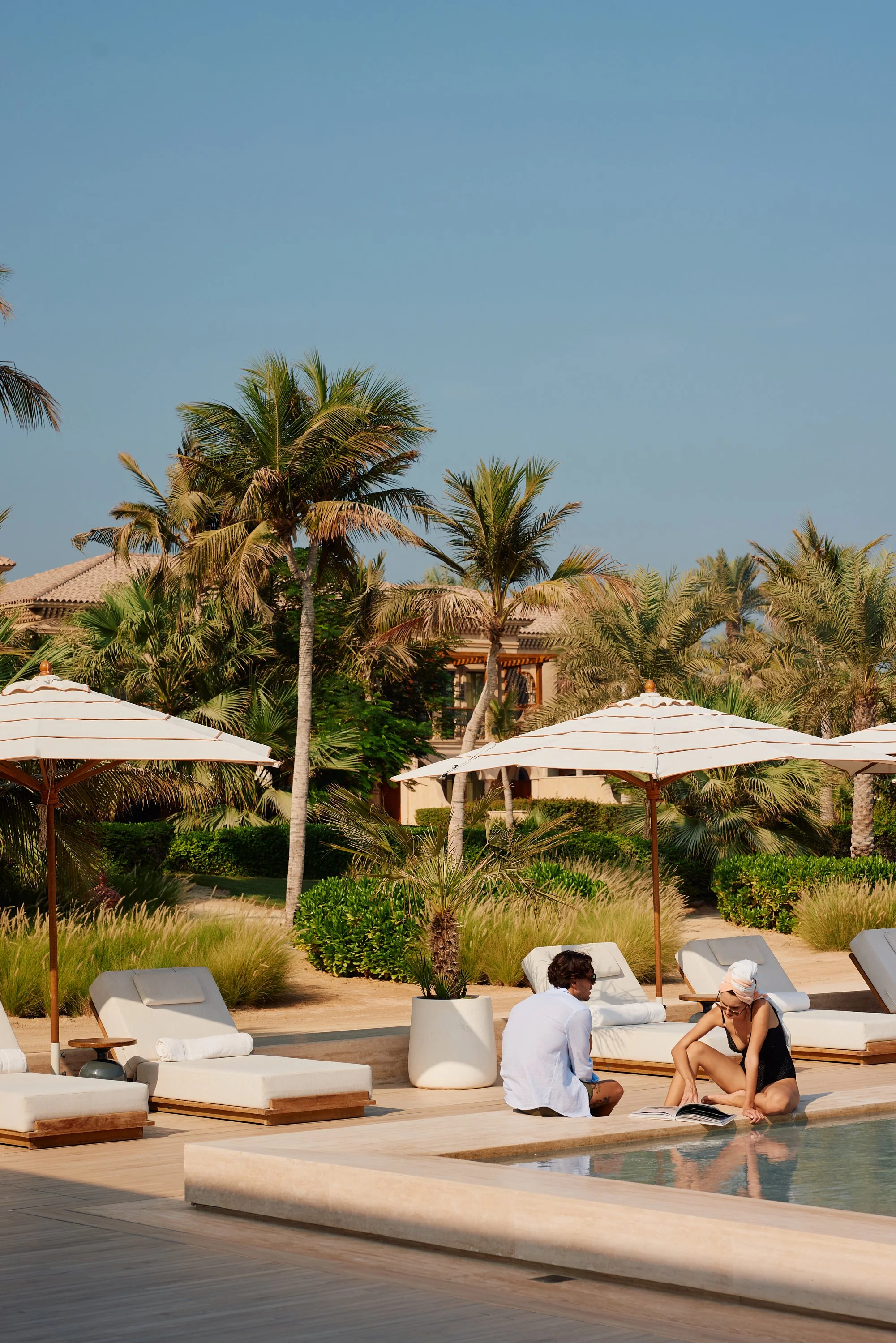 Two people sitting by a poolside under white umbrellas with palm trees and resort buildings in the background.