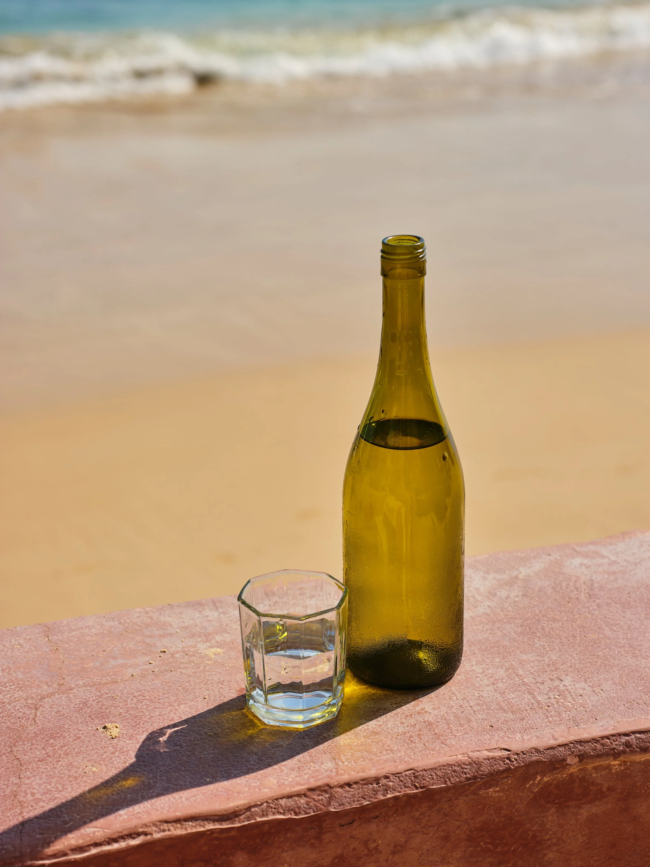 A green glass bottle and a clear glass with water are placed on a pink surface on the beach, with the ocean and waves in the background.