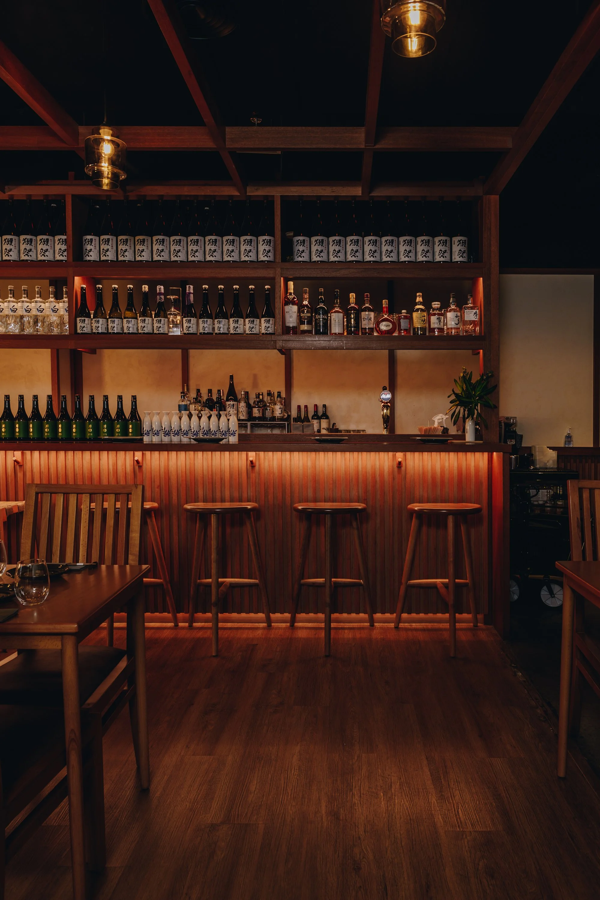Interior of a cozy bar with a backlit wooden counter, four stools, and shelves of liquor bottles, with warm lighting and wooden furnishings.