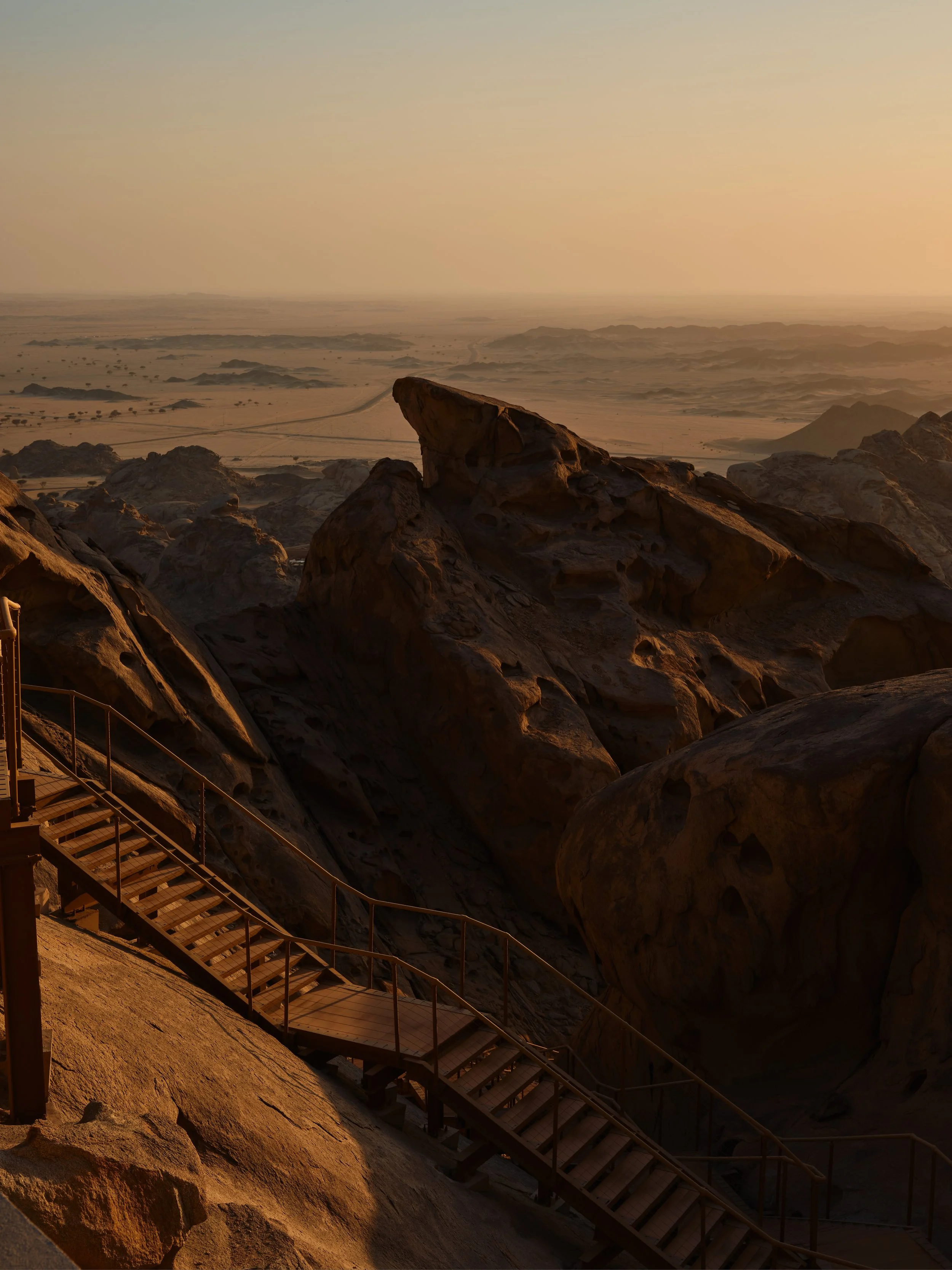 A desert landscape at sunset with large rock formations and a wooden staircase in the foreground.