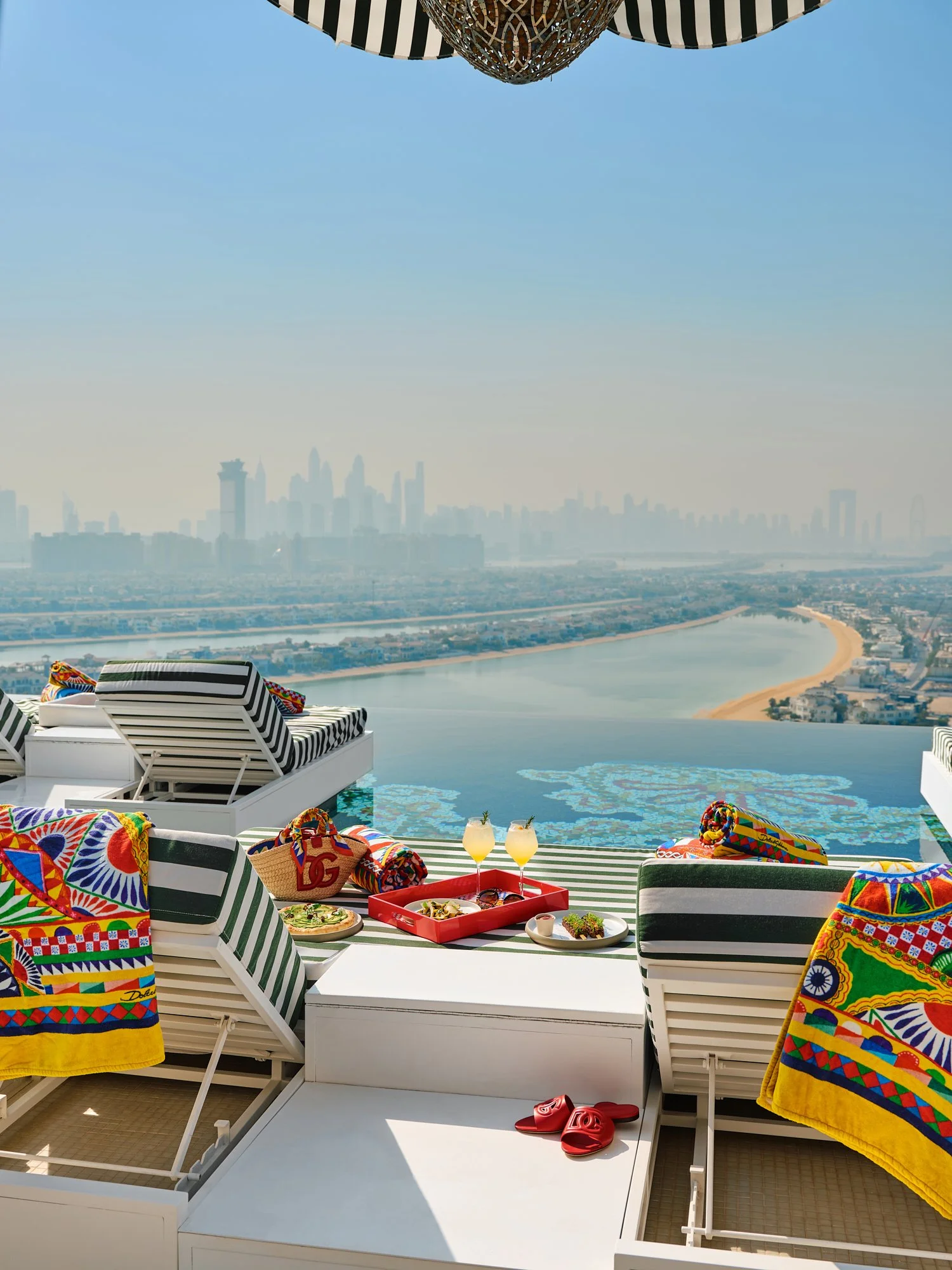 A rooftop infinity pool with a view of a city skyline, featuring beach towels, lounge chairs, a tray with drinks and food, and colorful towels and slippers.