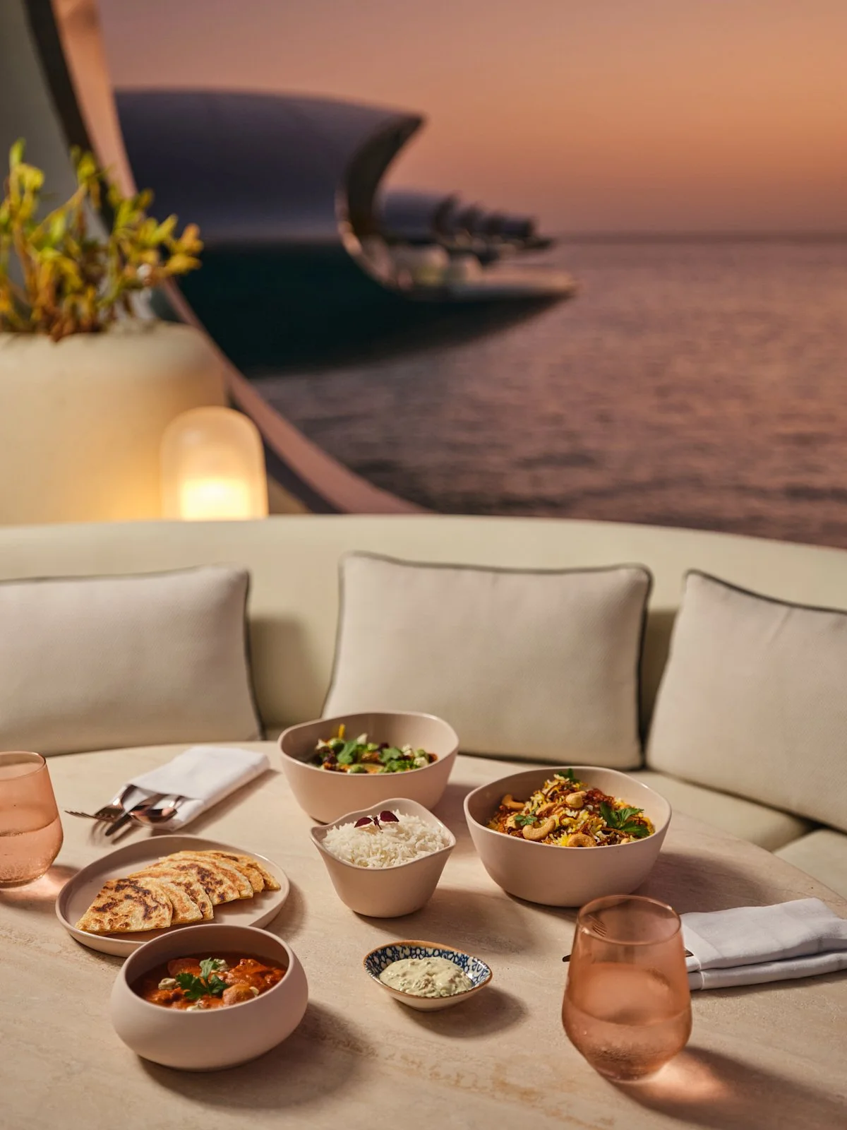 A dining table with Indian food dishes, including rice, curry, naan, and salad, set with pink glasses, white napkins, and silverware, with a seaside sunset mural in the background.