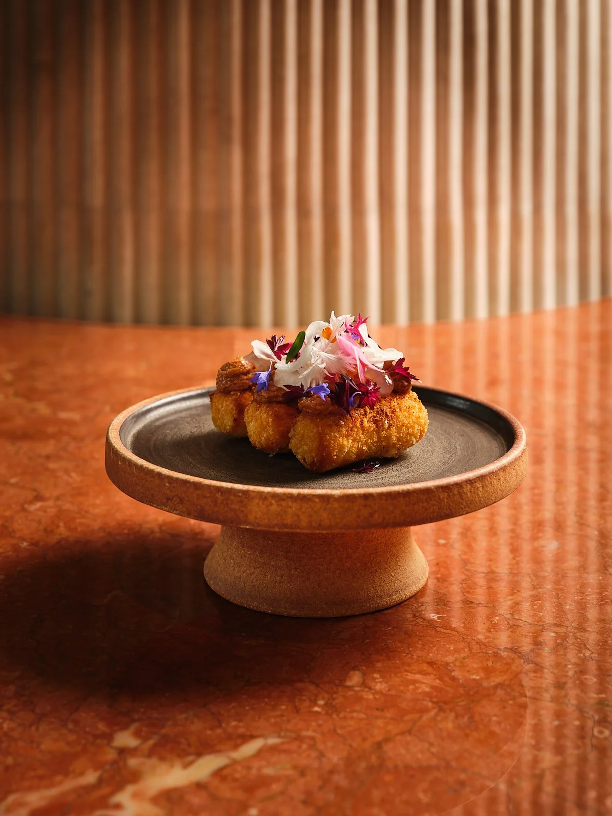Fried food garnished with edible flowers on a black ceramic plate, on a round wooden stand, with a textured background.