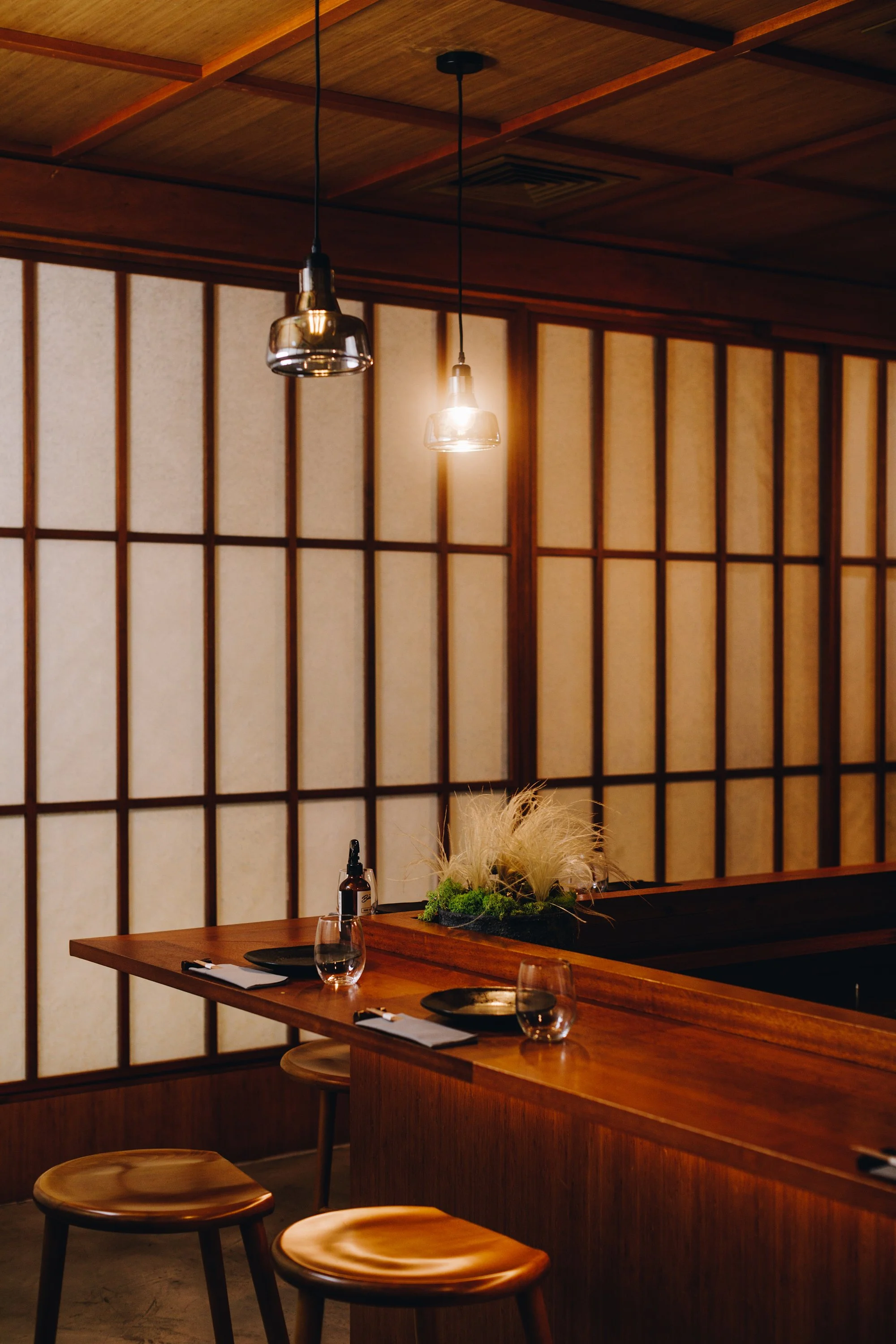 A cozy restaurant interior with a wooden bar, three hanging pendant lights, and traditional shoji-style wall panels. The bar is set with glasses, plates, a dropper bottle, and a decorative arrangement of plants and pampas grass.