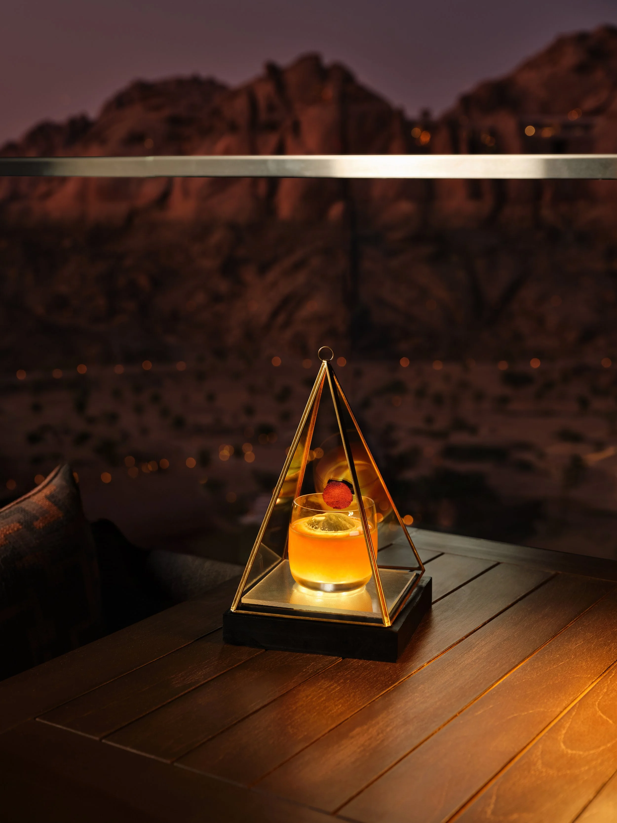 A drink with a red cherry garnish in a glass, placed inside a gold triangular frame on a wooden table against a mountainous landscape at night.