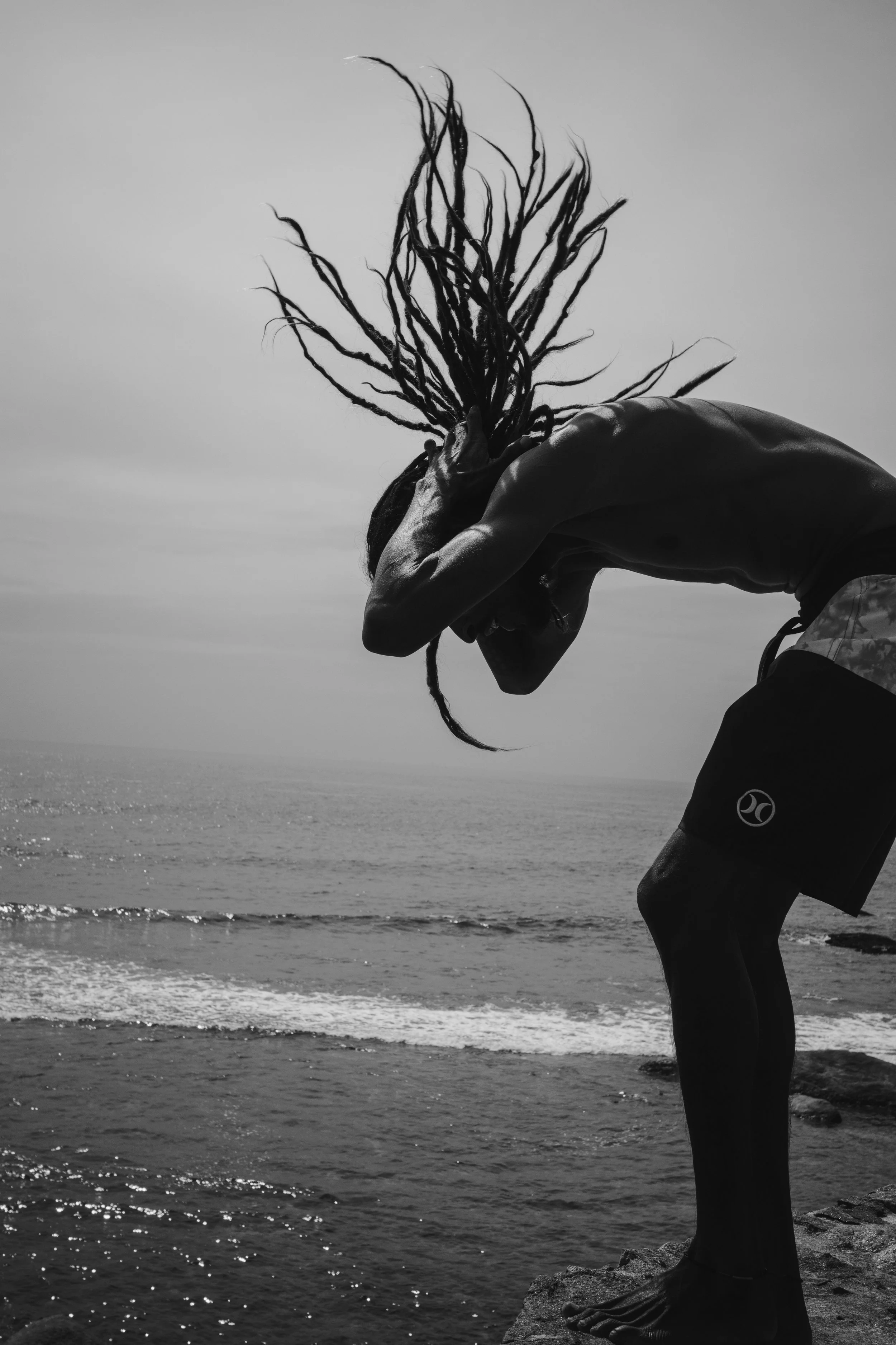 A silhouette of a person with dreadlocks, bending over near the water, on a beach or rocky shoreline, against a cloudy sky.