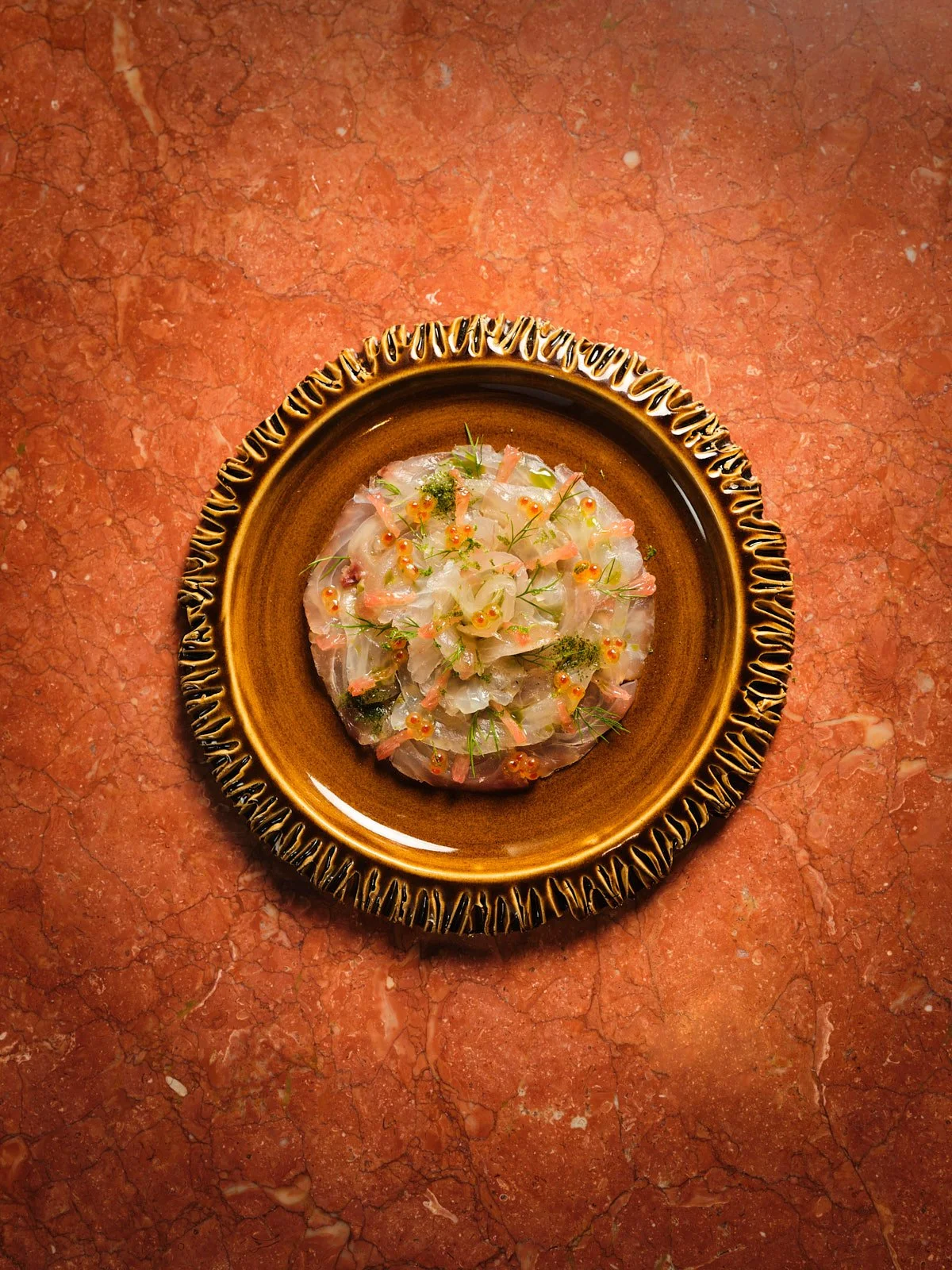 Thinly sliced raw fish, garnished with herbs, dots, and microgreens, arranged in a circular pattern on a brown plate with ridged edges, on a red marble surface.