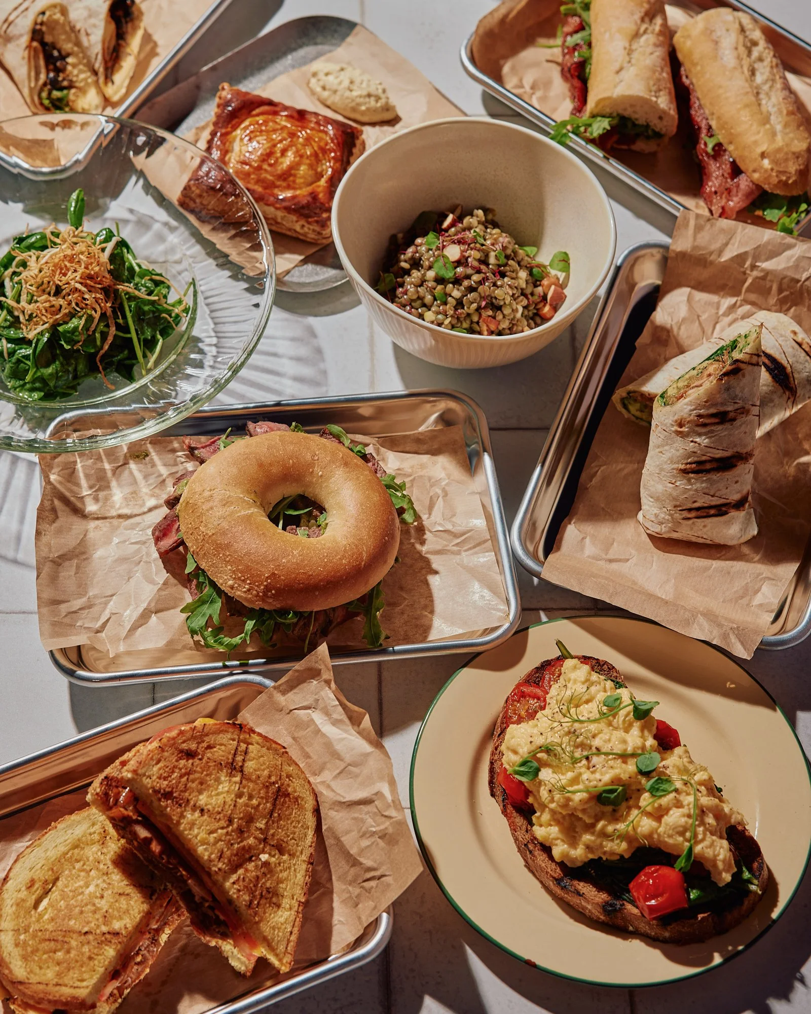 A variety of sandwiches, salads, and baked goods on a table, including a bagel sandwich, wraps, a grilled sandwich, a side salad, and a small bowl of grains with vegetables.
