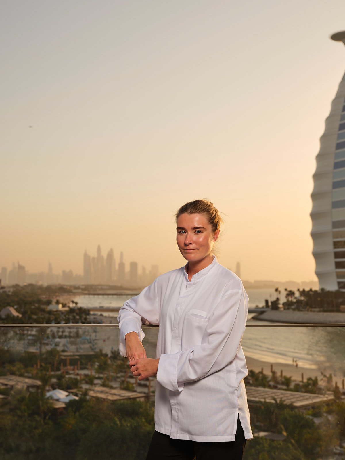 A woman in a white chef's coat standing outdoors on a balcony with a city skyline and beach in the background during sunset.
