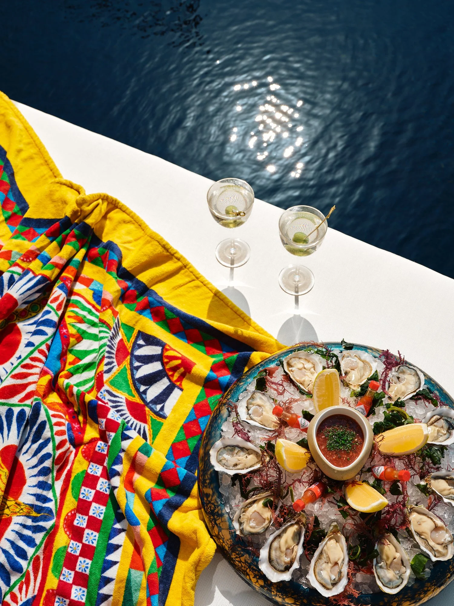 Plate of oysters with lemon wedges and cocktail sauces, two glasses of cocktails with olives, colorful cloth, and ocean water in the background.