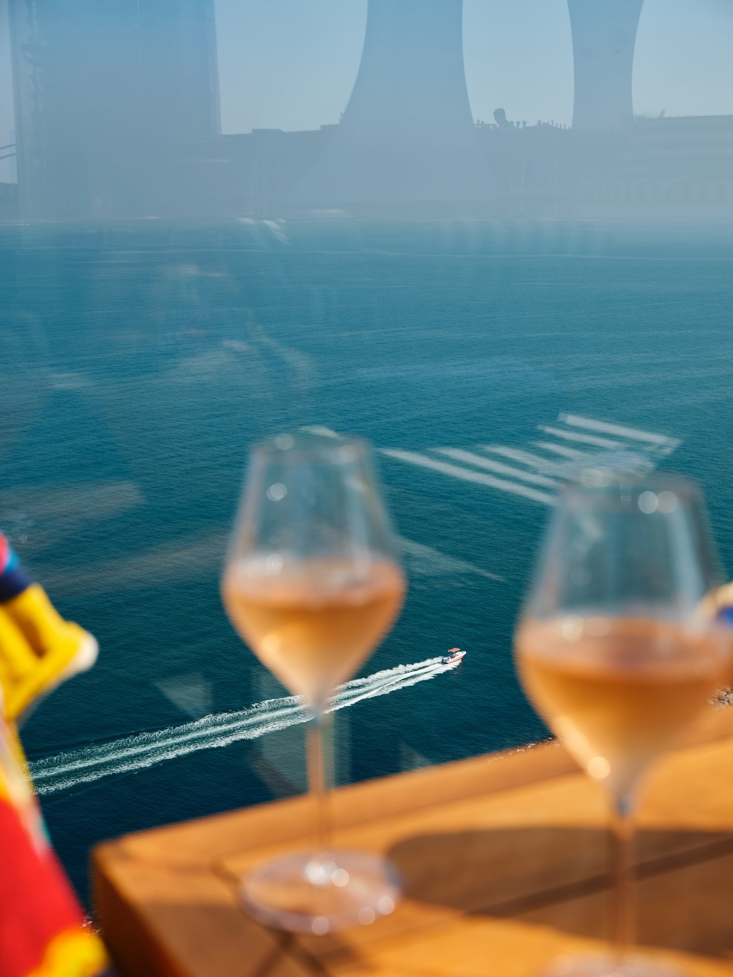 View of the ocean with a boat moving across, seen through a window reflection. Two glasses of rosé wine on a table in foreground.