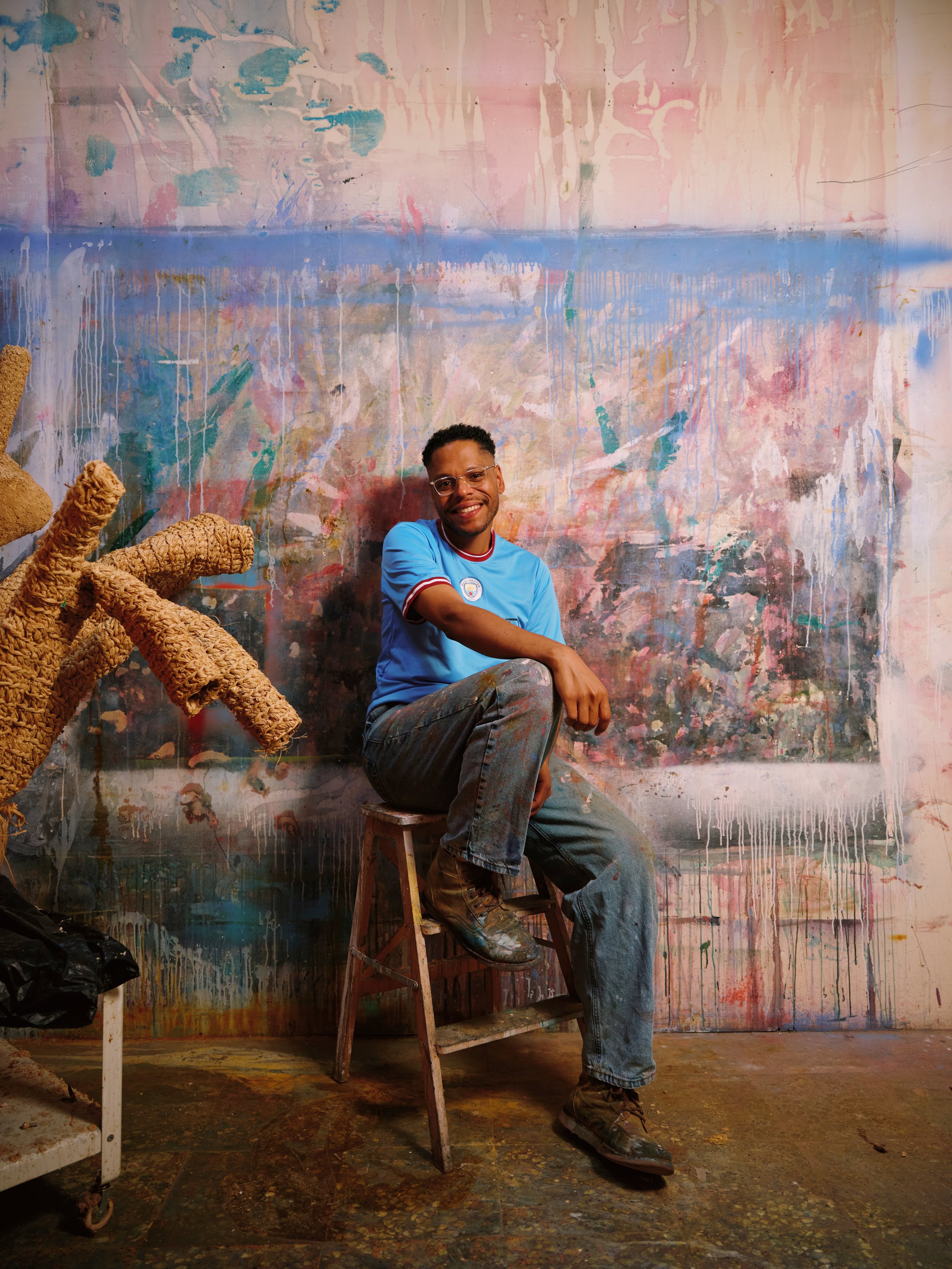 A young man sits on a step ladder in front of an abstract, colorful wall with drips and textured paint. He is smiling, wearing glasses, a blue shirt with a badge, and jeans with paint stains. There are two artistic textile sculptures on the left side of the image.