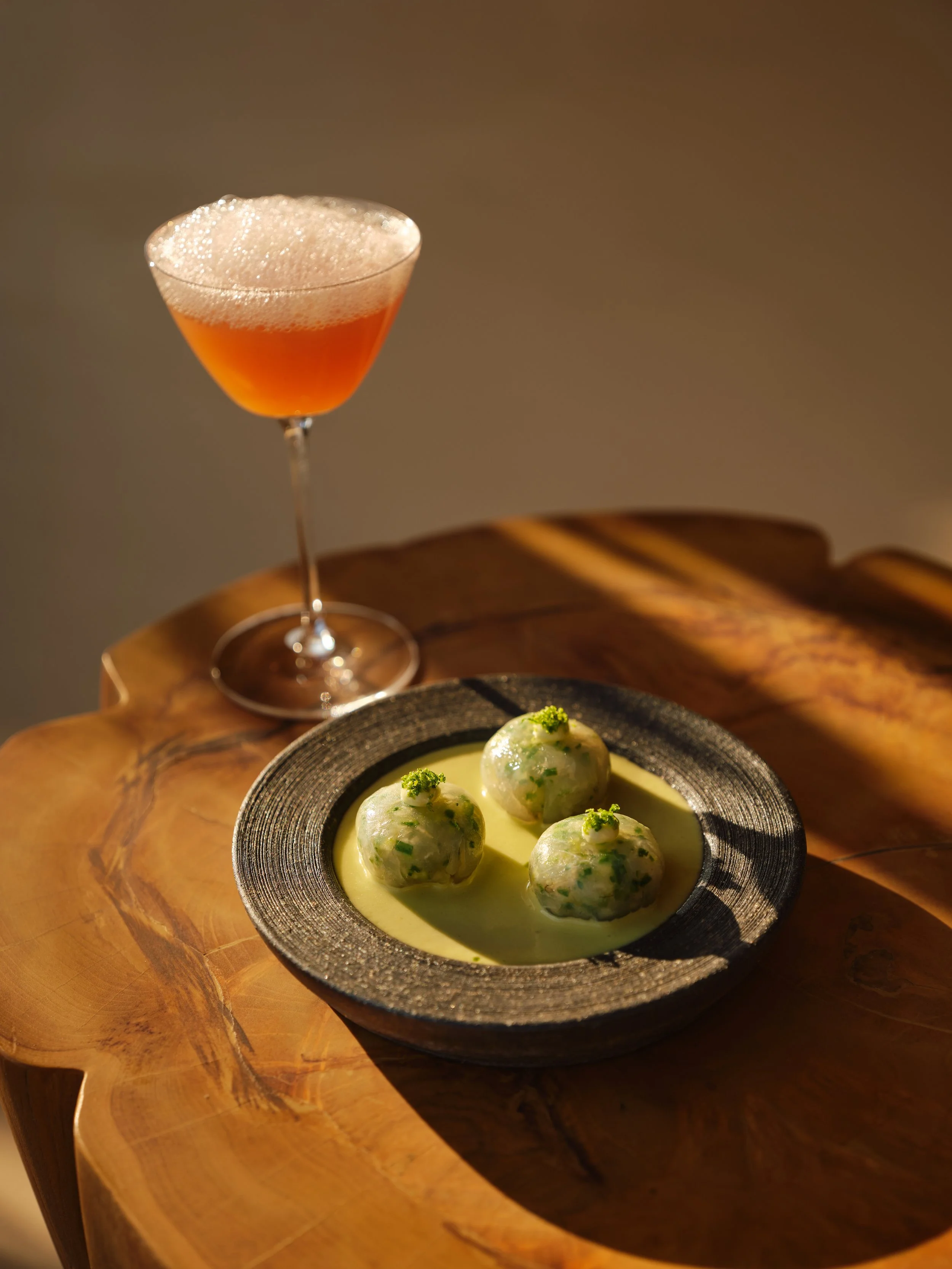 A cocktail in a martini glass and a black plate with three greenish meatballs garnished with chopped herbs and served in a creamy yellow sauce, placed on a wooden table.