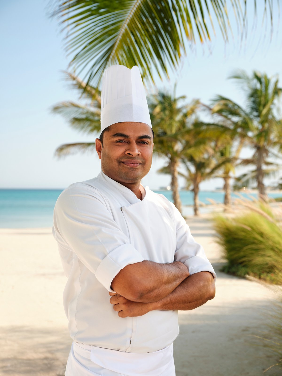 A male chef in a white uniform and tall chef's hat stands on a beach with palm trees in the background, crossing his arms and smiling.