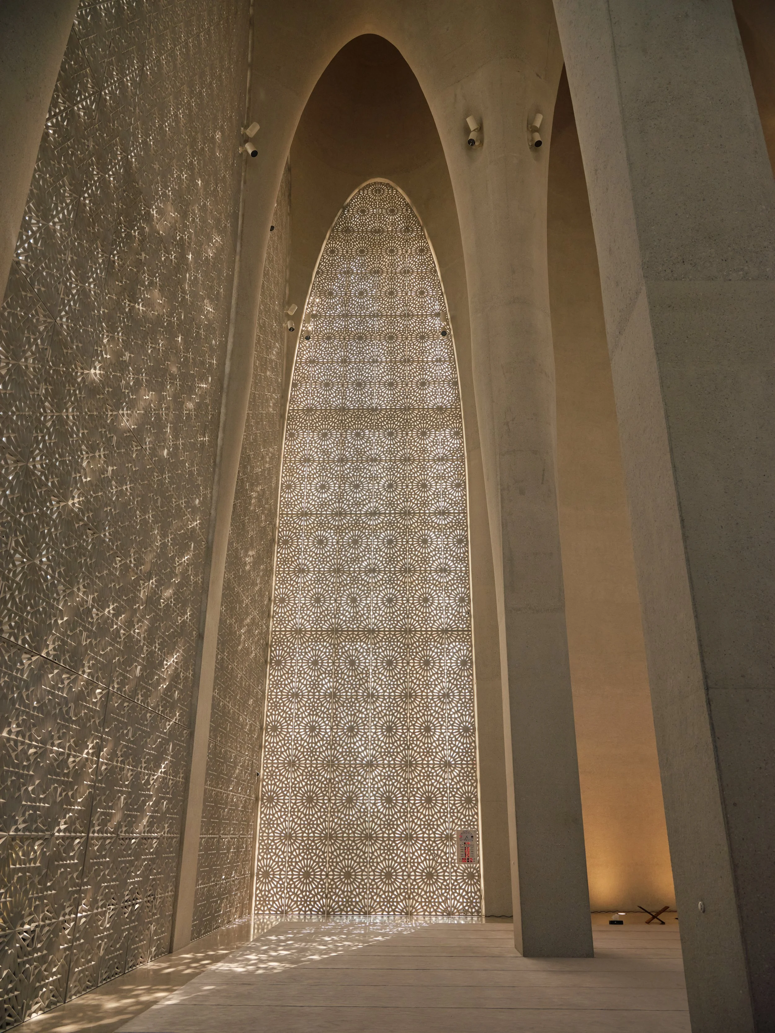 A tall, arched window with intricate geometric lattice patterns allowing light to cast decorative shadows inside a modern interior with concrete walls and columns.
