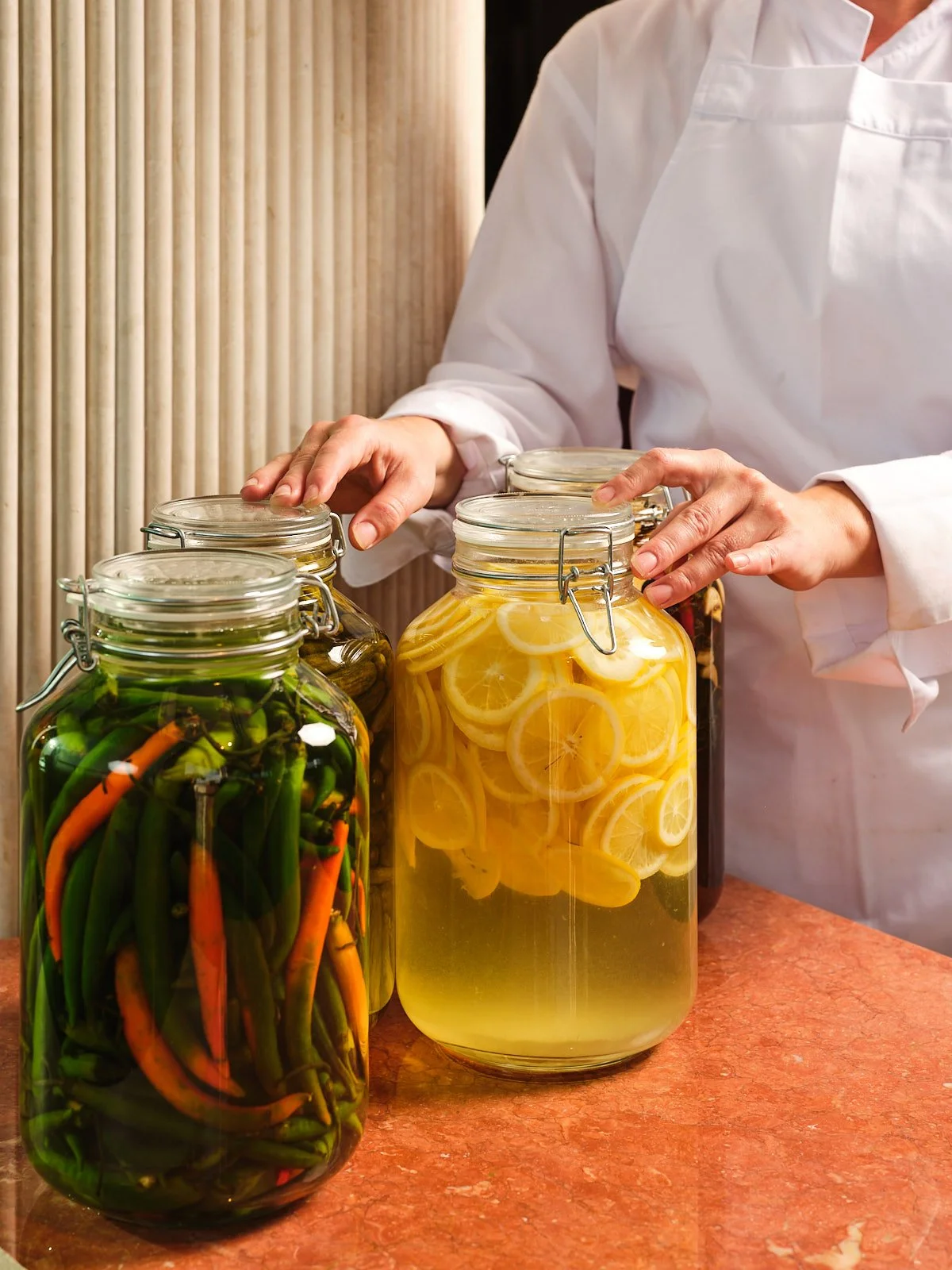 Person in white coat arranging jars of homemade pickled vegetables and lemon-infused water on a kitchen counter.