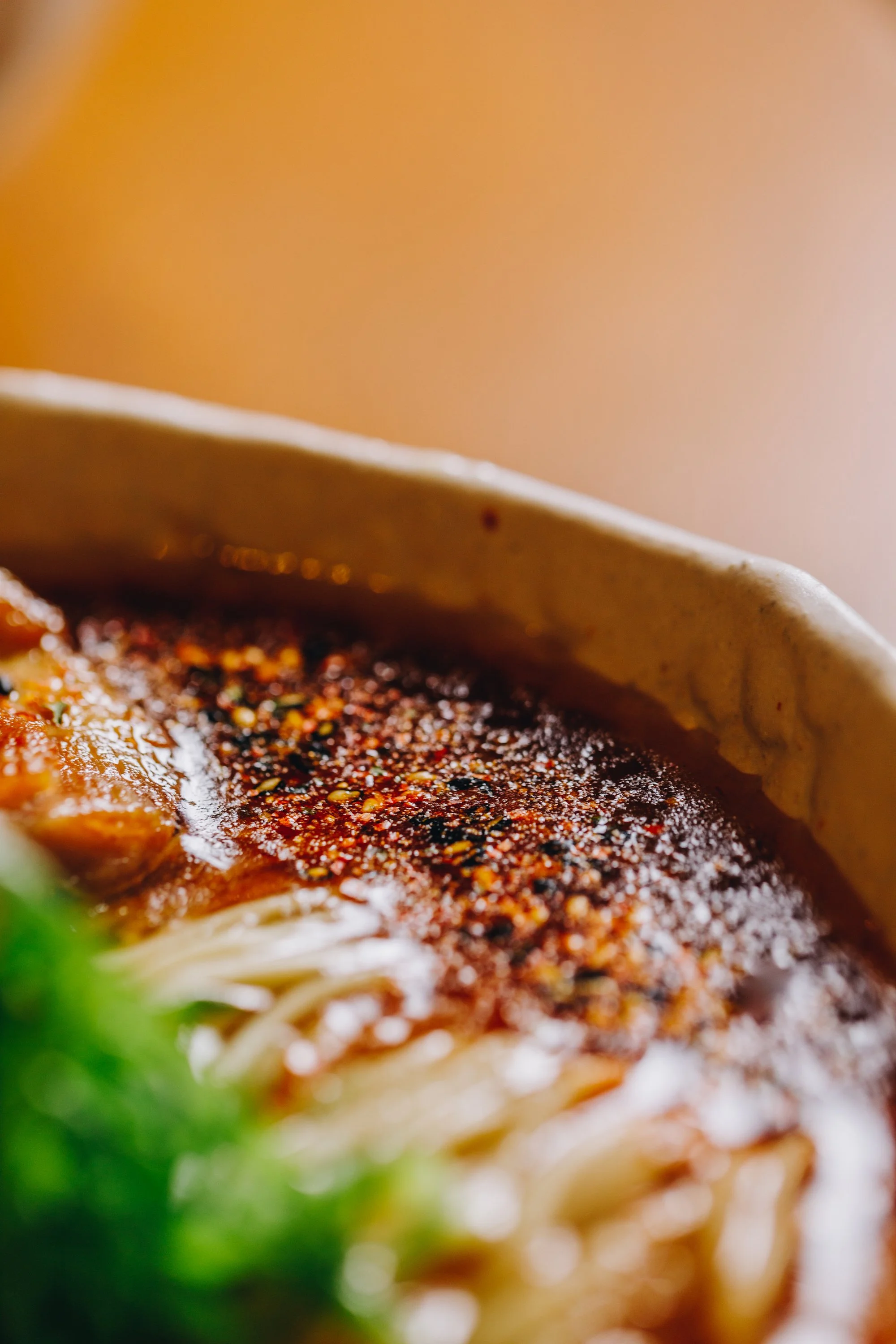 Close-up of a bowl of ramen noodles with red chili oil and pepper flakes on top, green vegetables partially visible in the foreground.