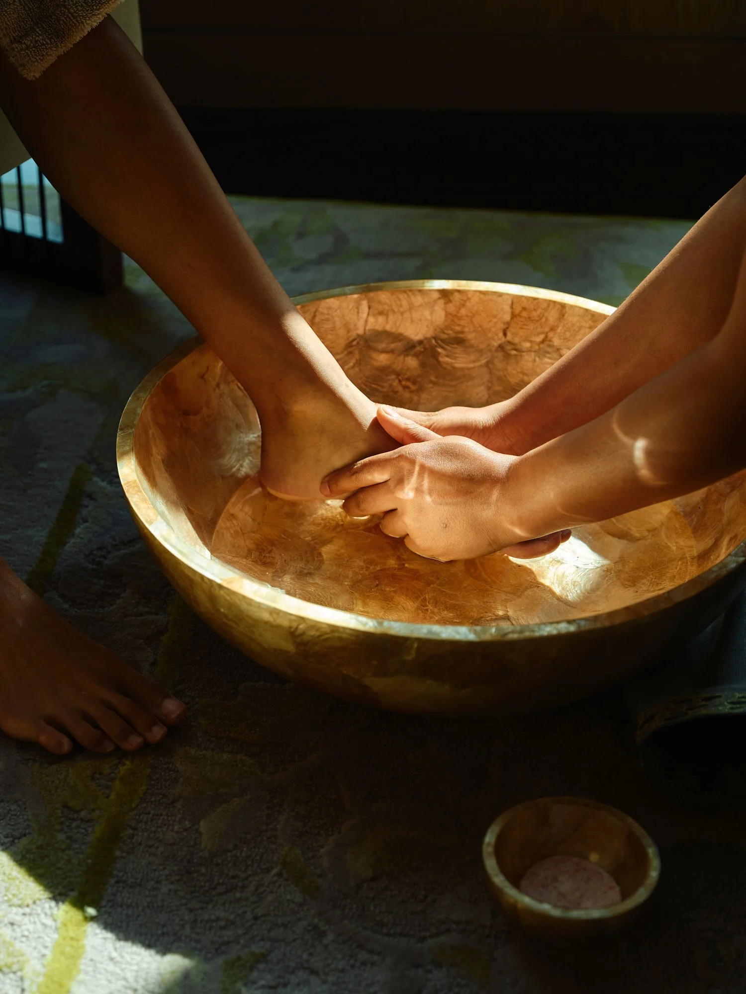 Two people soaking their feet in a wooden bowl filled with water, with sunlight casting shadows.