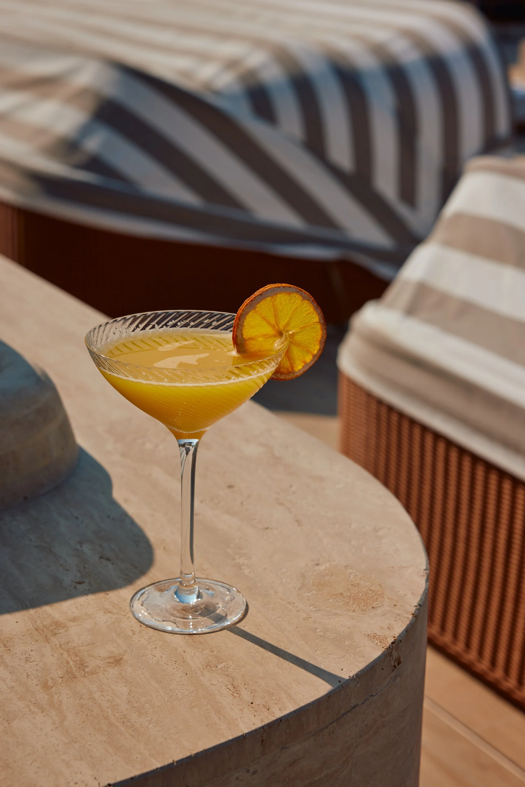 A yellow cocktail in a coupe glass with a dried orange slice garnish, placed on a light-colored, rounded stone table.