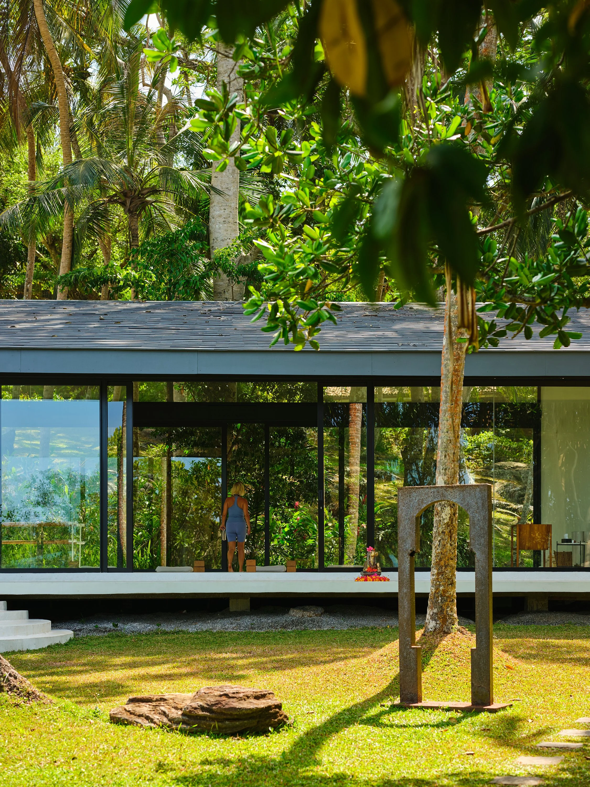 Modern glass house surrounded by lush tropical greenery, with a woman in a blue outfit standing inside, and a rock with a metal sculpture in the foreground.