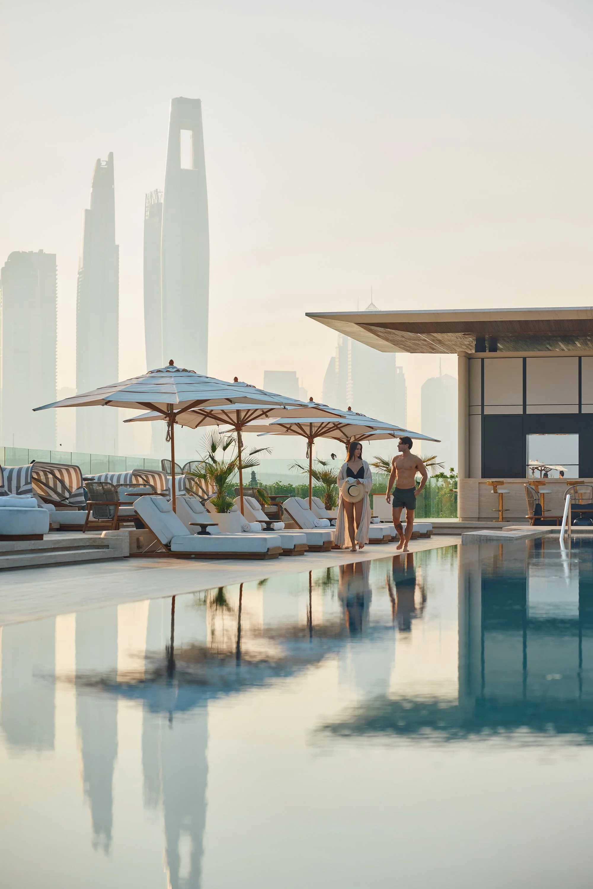 A rooftop pool area with lounge chairs and umbrellas, two people walking near the pool, city skyline in the background.