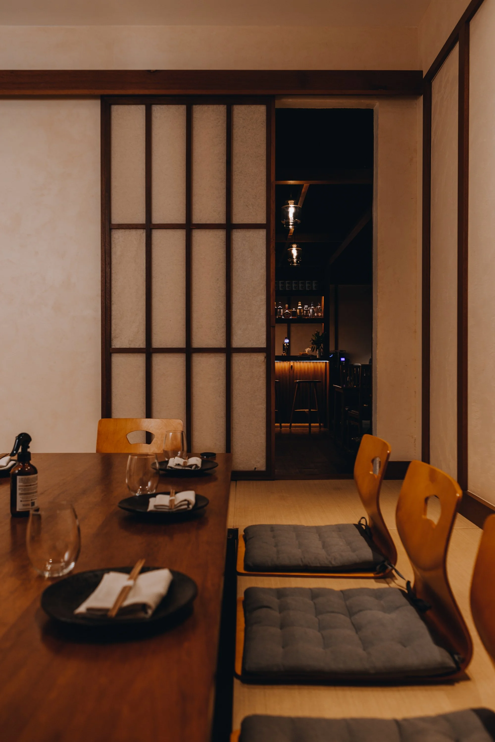 A cozy Japanese-style dining room with a wooden table set with black plates, chopsticks, glasses, and a bottle. There are three wooden chairs with gray cushions, and a sliding shoji screen separates the dining area from a dimly-lit bar area in the background.